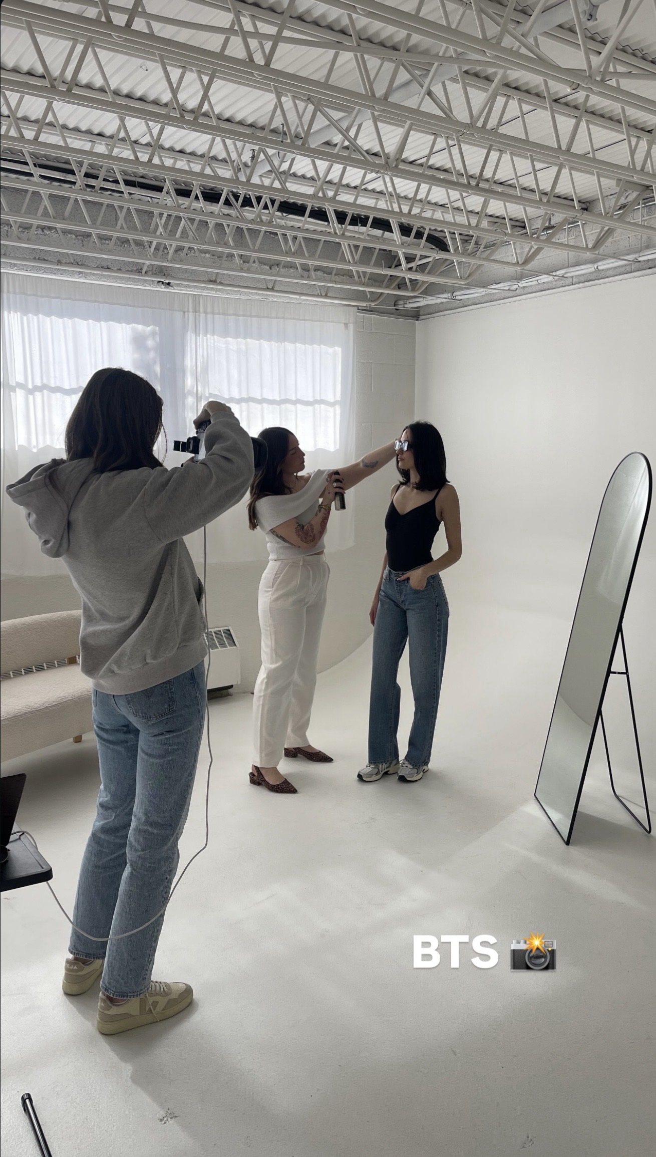 Two women are posing for a photo in a studio, with a photographer taking their picture. The woman on the right is wearing a black tank top and jeans, and the woman on the left is adjusting her sunglasses. There is a large mirror on the right side and a white couch in the background.