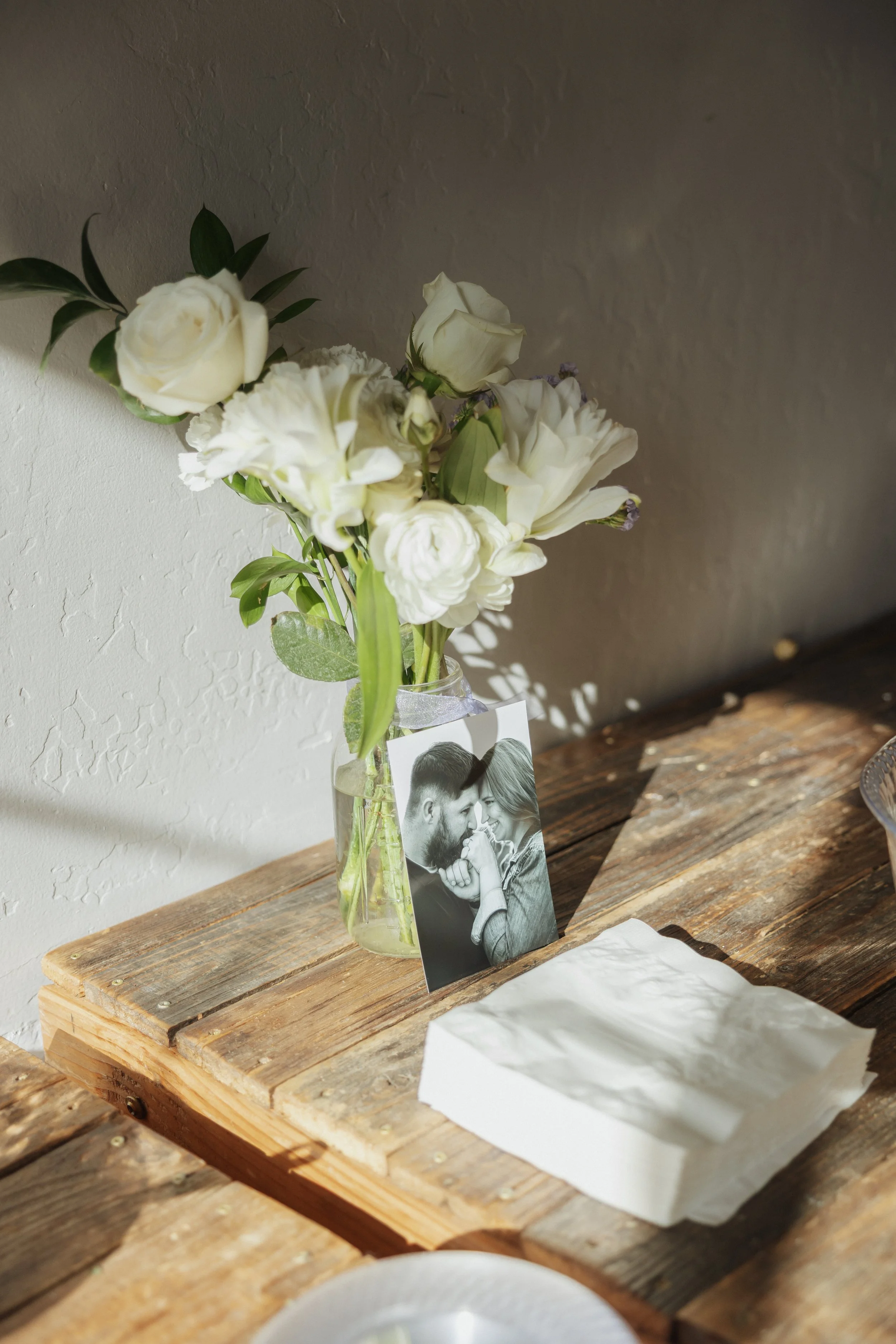 A bouquet of white flowers in a glass vase placed on a rustic wooden table, with a black and white photograph of a couple leaning close and smiling at each other underneath the vase.