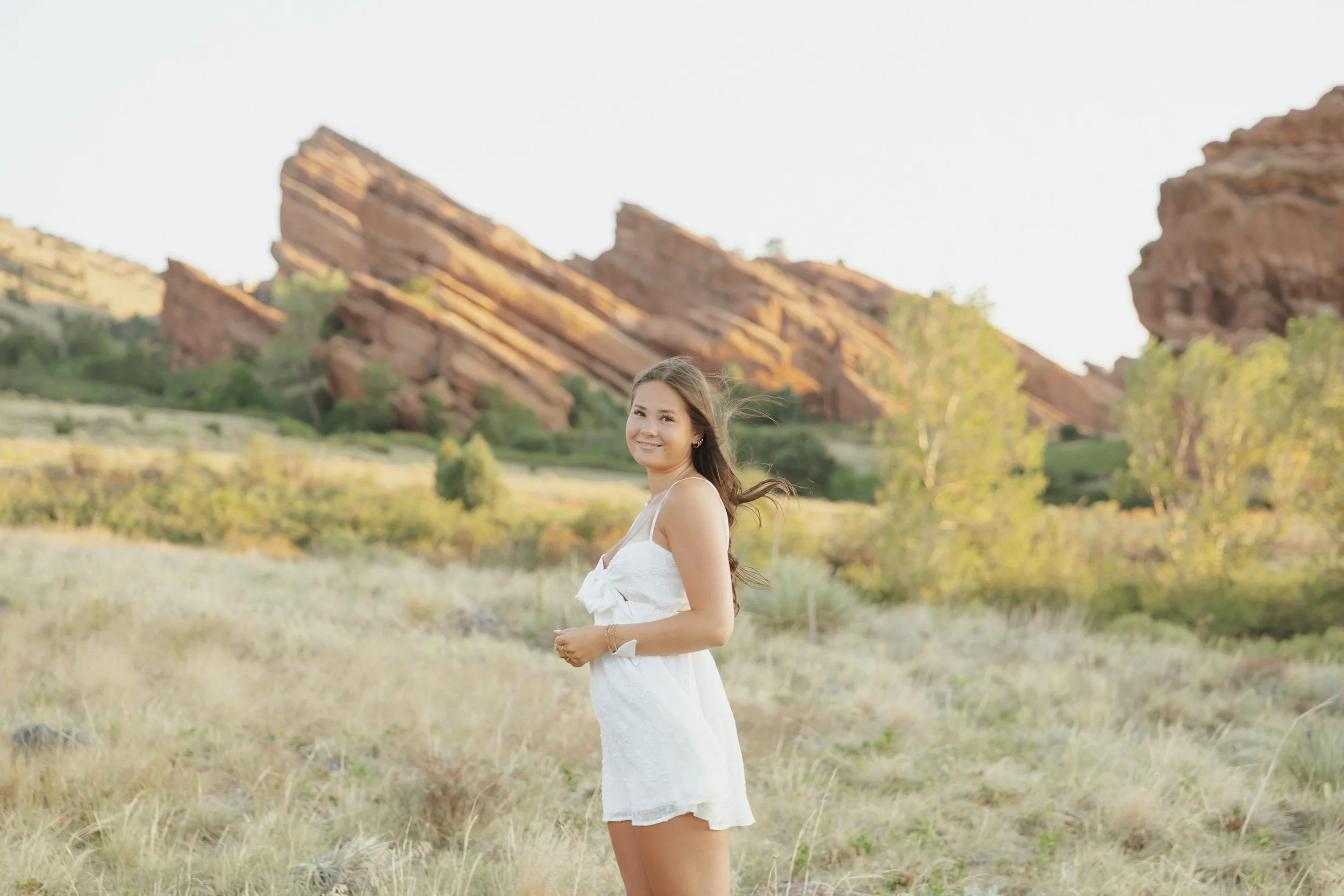 A young woman in a white dress standing in a field with grass and small plants, smiling, with large red rocks or cliffs in the background and a clear sky.