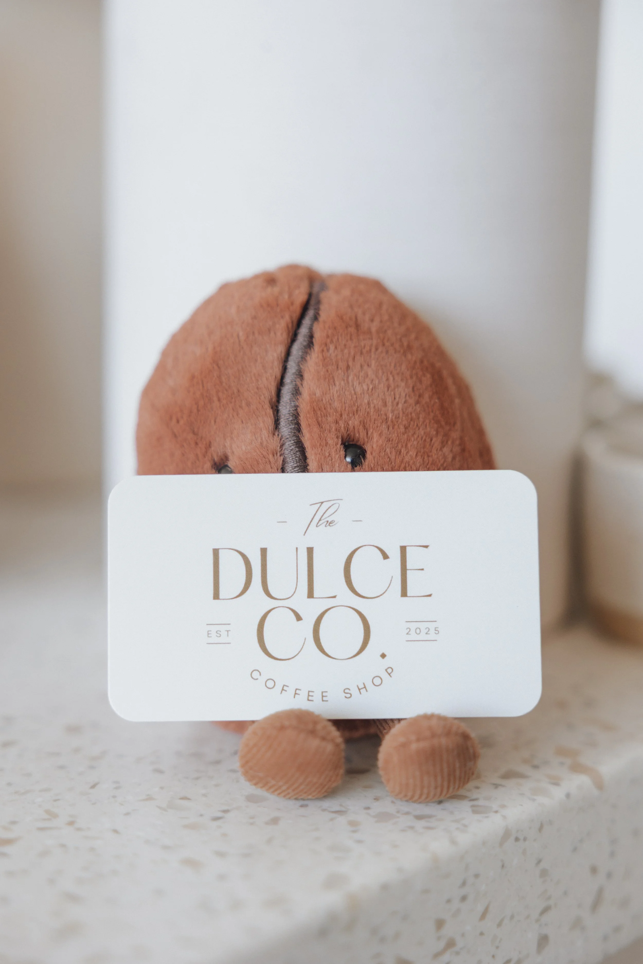 A close-up of a plush coffee cup with a lid, standing behind a restaurant menu card that reads 'The Dulce Co. Coffee Shop 2025' and shows an image of a brown plush coffee cup with a black line running down the middle, on a speckled countertop.
