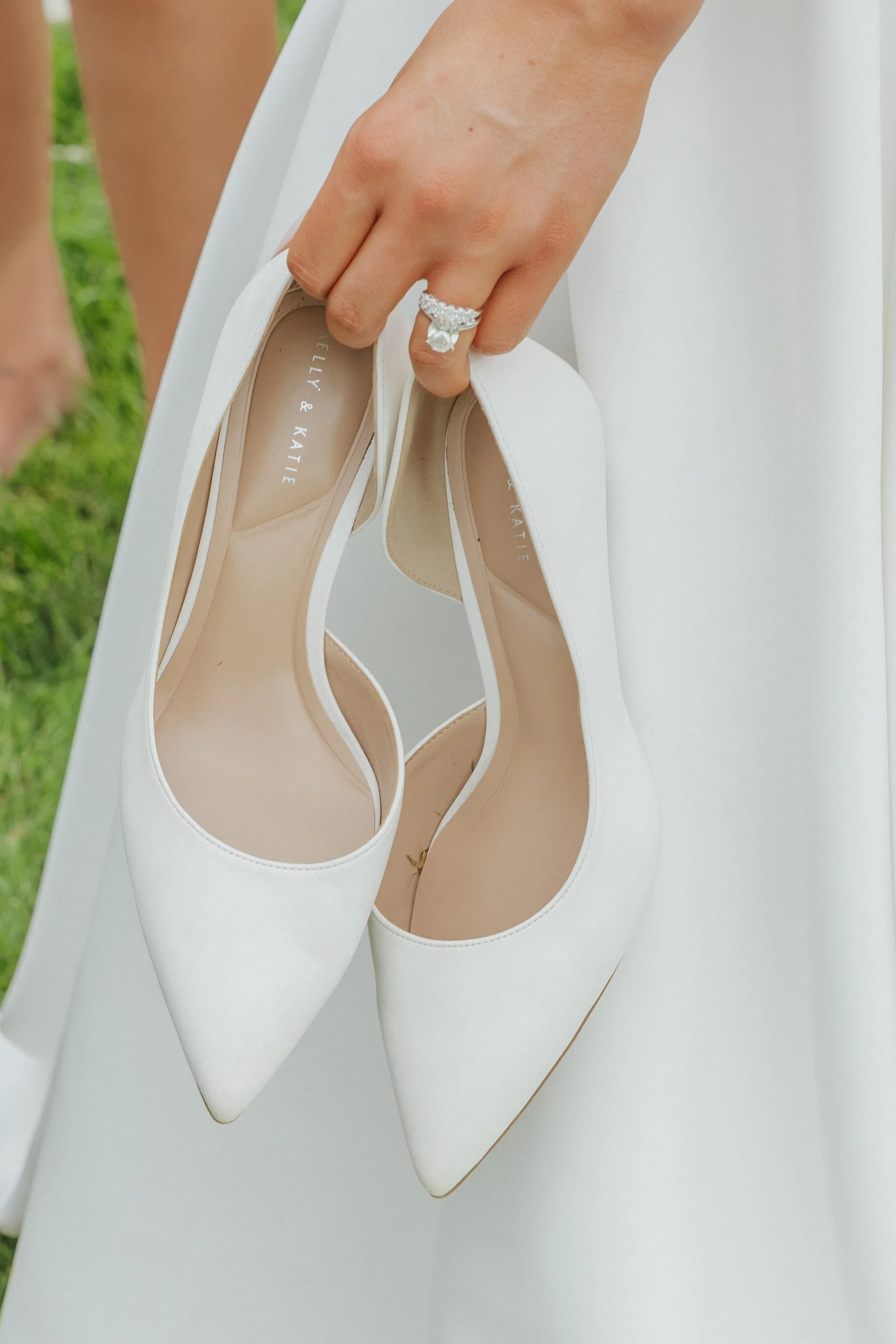 A hand holding a pair of white high-heeled shoes, with a large diamond ring on the index finger. The shoes are labeled with the brand "BELLY & KATIE" and are resting on a white surface. The background is green and out of focus.