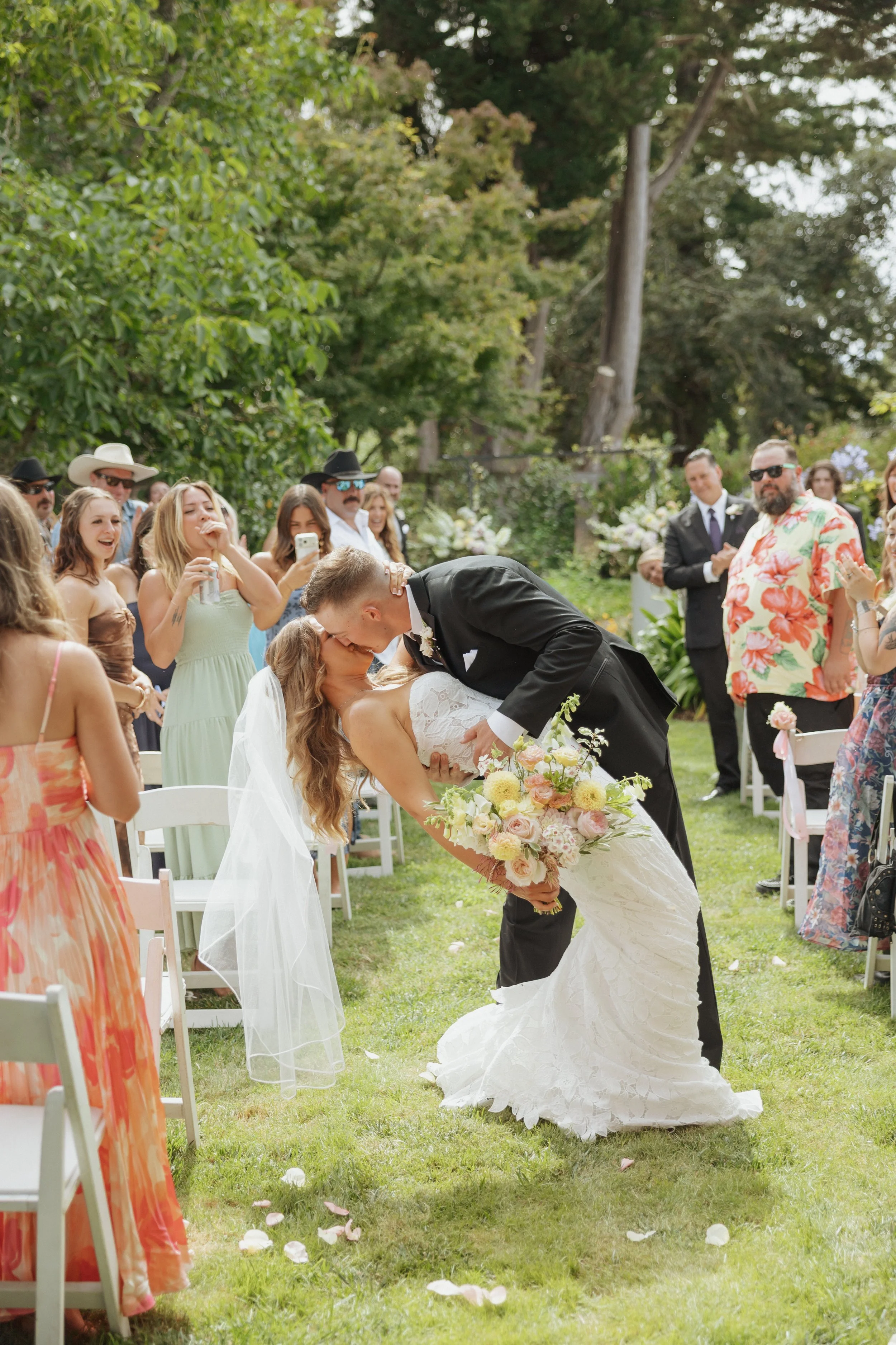 A newlywed couple sharing a kiss during their outdoor wedding ceremony, surrounded by friends and family, with lush green trees in the background and floral decorations.