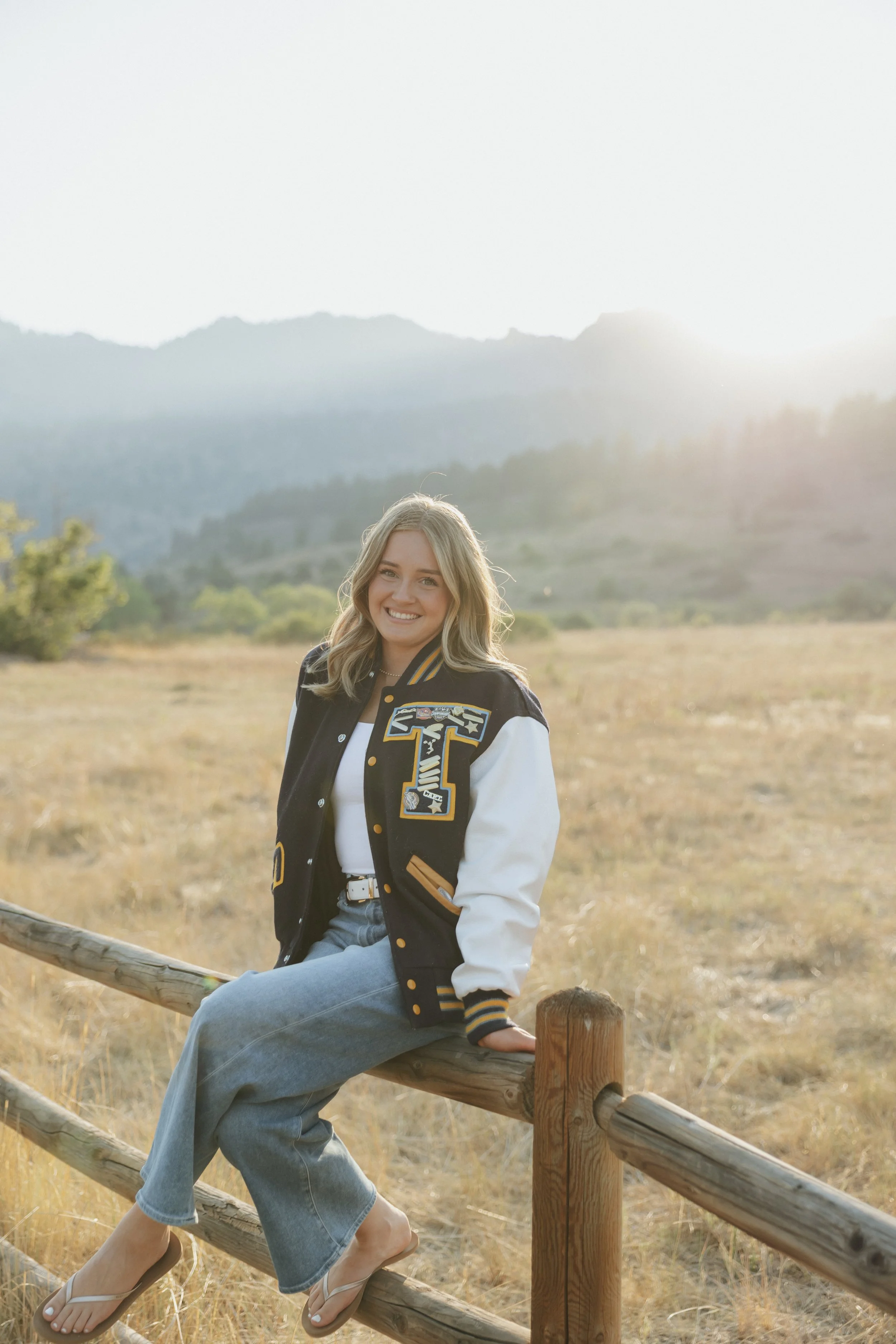 A smiling young woman with blonde hair sitting on a wooden fence in an open field with mountains in the background during sunset.