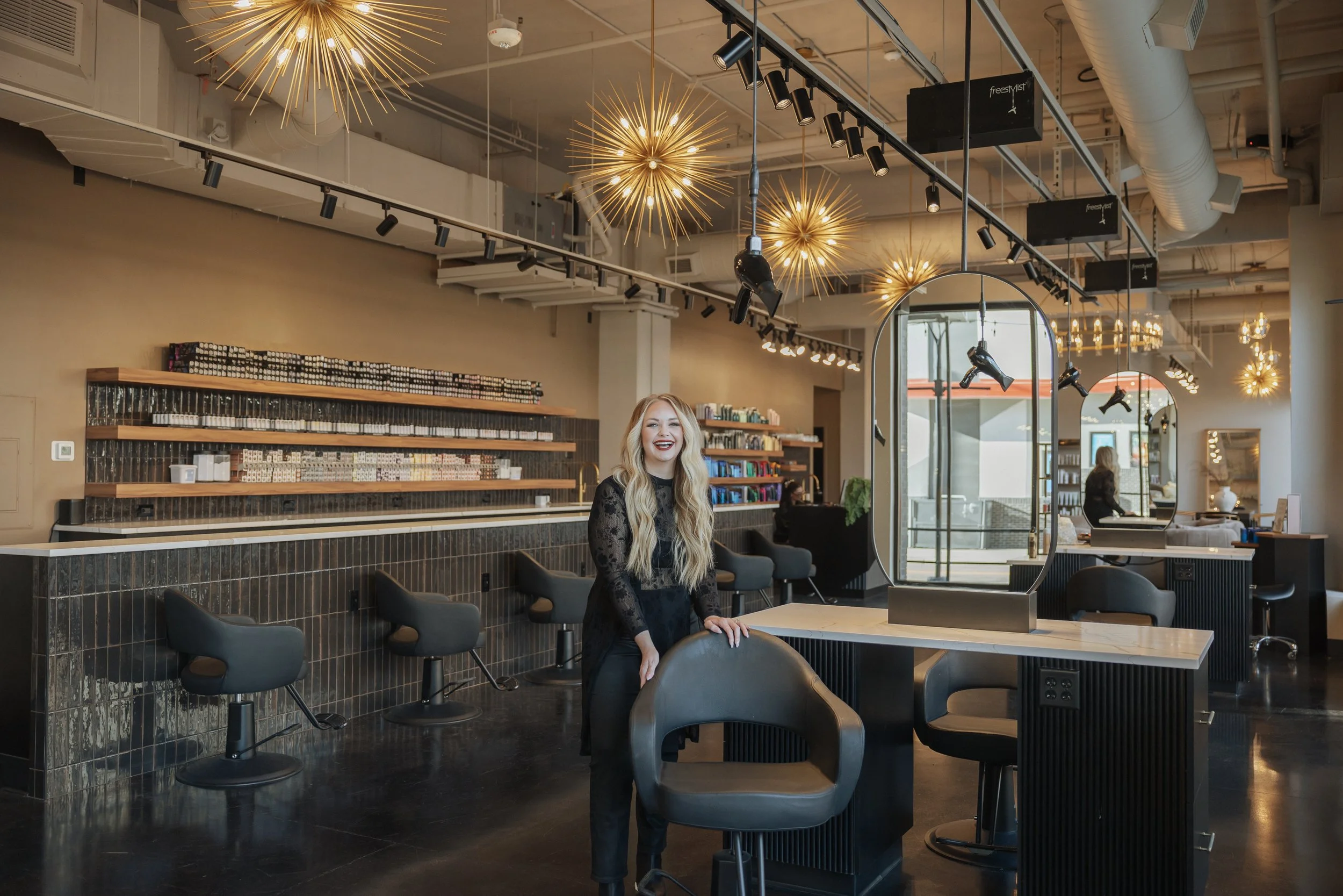 A woman with long blonde hair wearing a black lace dress, smiling, standing in a modern salon or spa with hanging pendant lights, mirrors, and shelves filled with products.