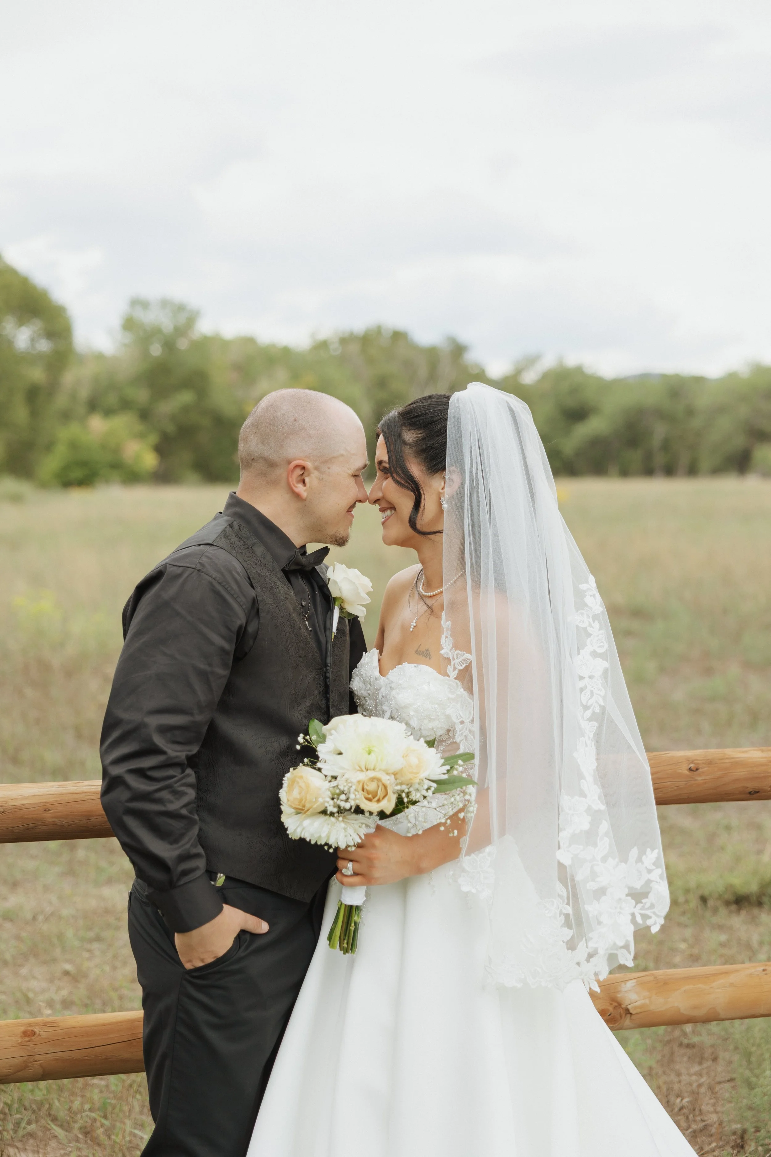 A groom and bride face each other with their foreheads touching, outdoors in a field with green trees in the background. The groom is bald, wearing a black shirt with a white boutonniere. The bride has dark hair, wears a white lace veil, strapless wedding gown, and pearl earrings. She holds a bouquet of white roses and cream-colored flowers.