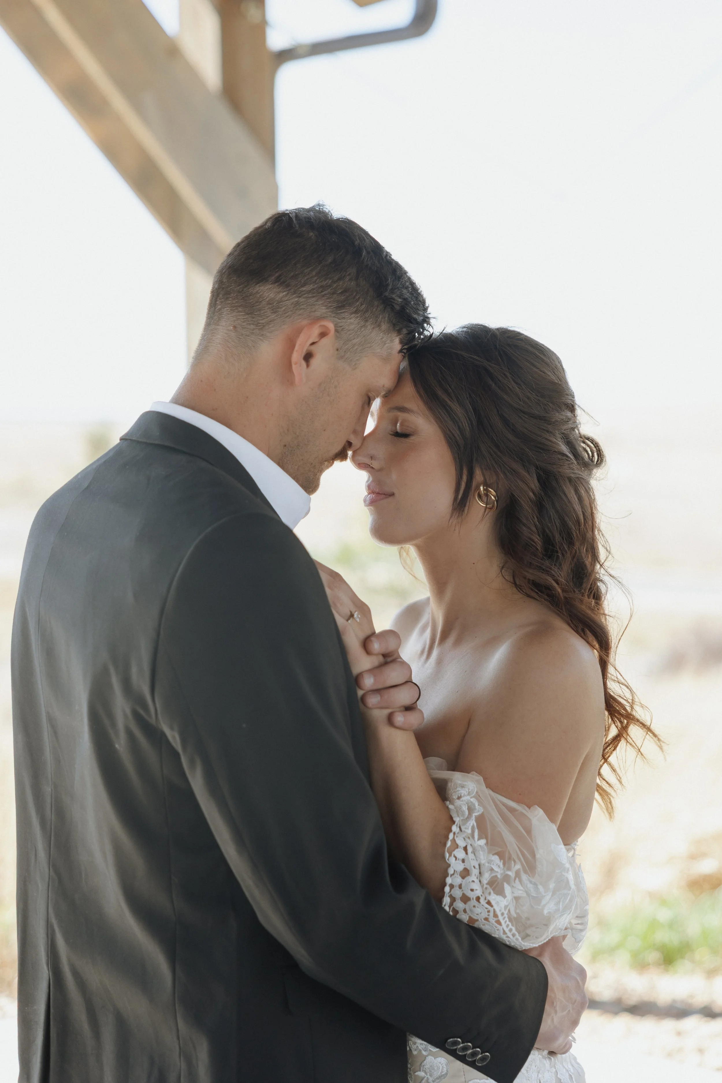 A bride and groom with foreheads touching, eyes closed, standing close and holding hands in a light-filled outdoor setting.