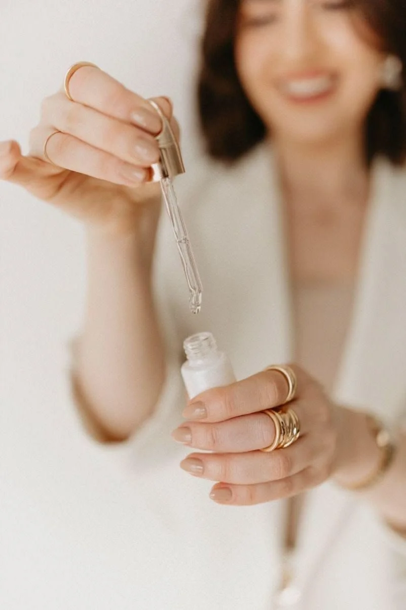 A woman with short dark hair and a white top is holding a small bottle of nail polish and using a dropper to put nail treatment onto her nails. She is wearing multiple rings on her fingers.