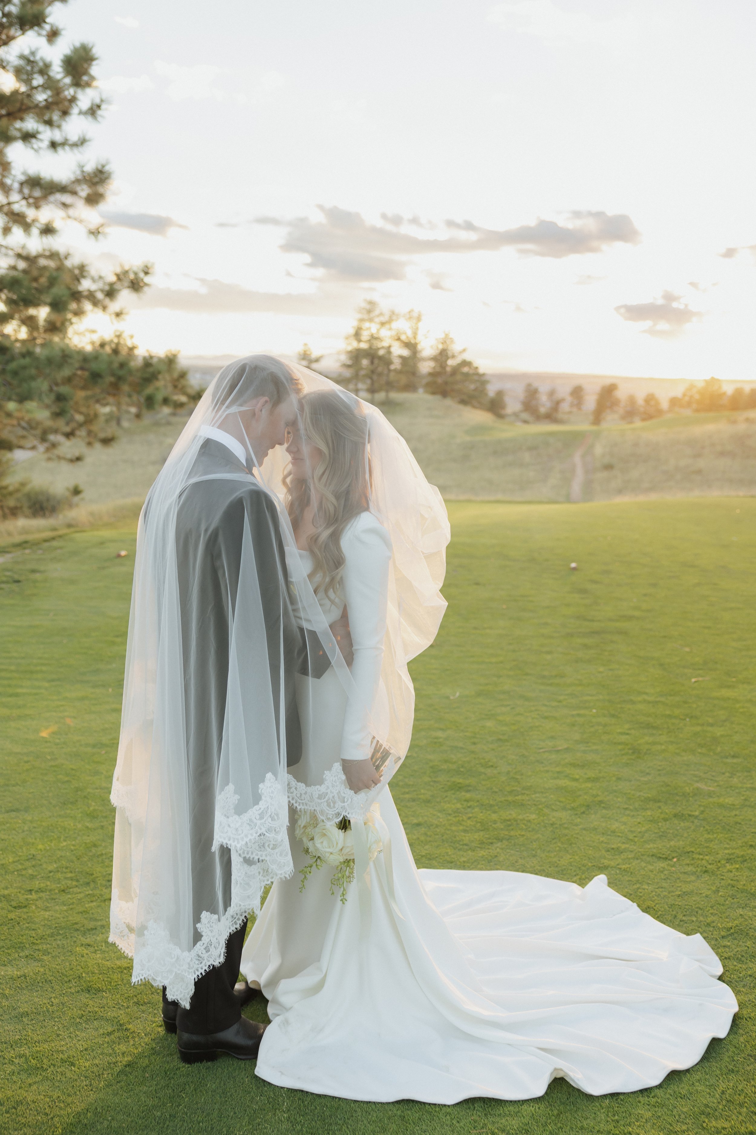 A bride and groom standing close together on a grassy field during sunset, with trees and a trail in the background, sharing a tender moment under a white veil.