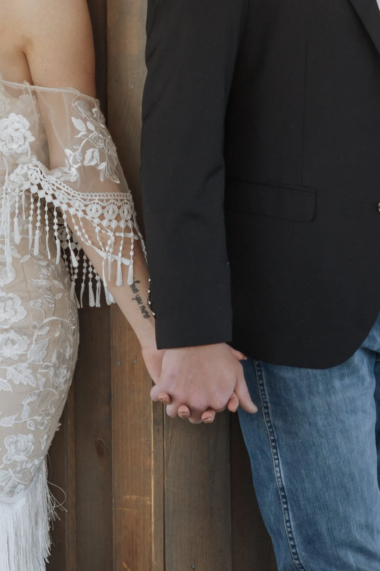 A couple holding hands, with the woman wearing a white lace dress and the man dressed in black and blue jeans, standing close together against a wooden background.