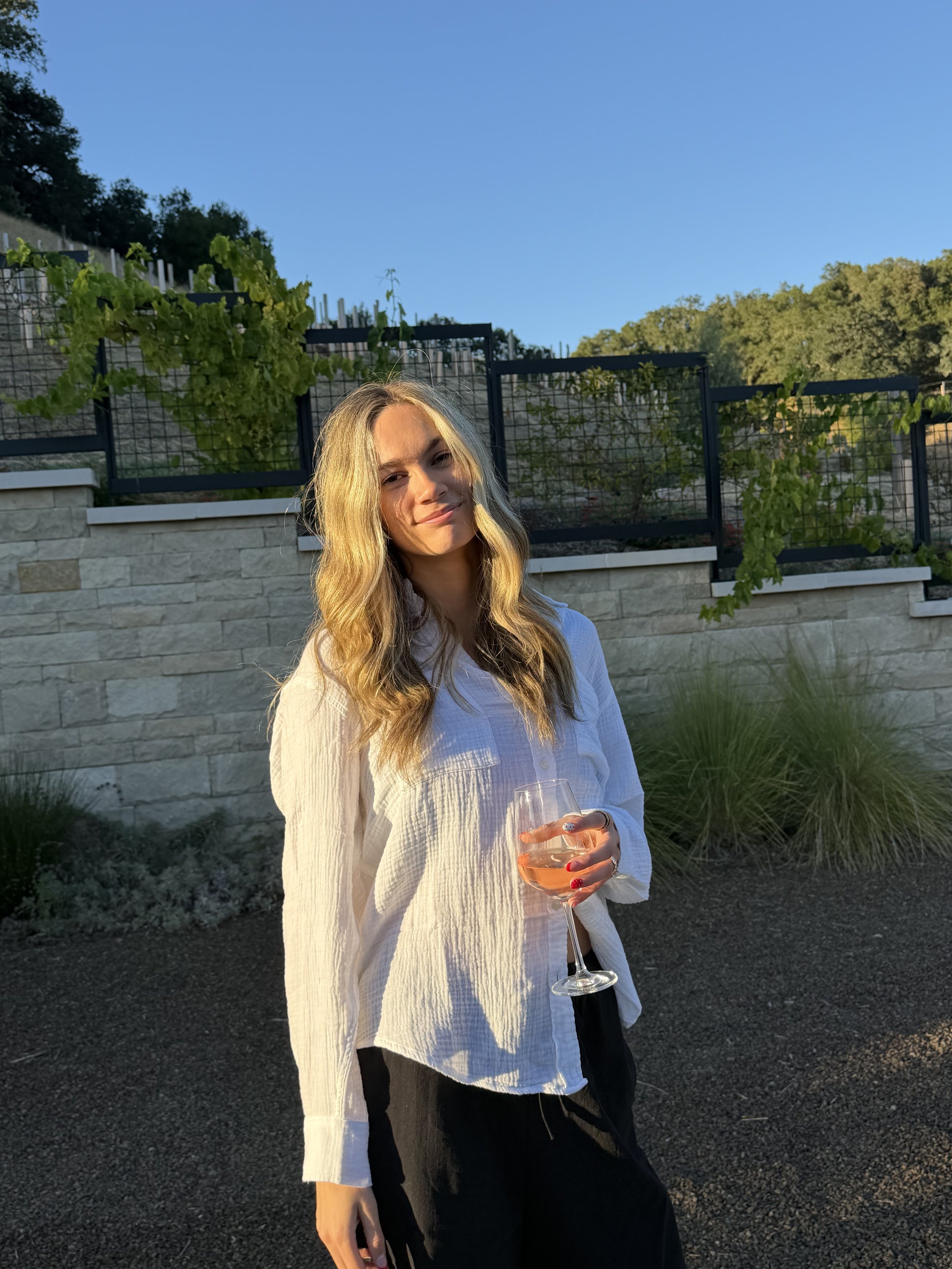 A woman with long blonde hair, wearing a white blouse and black pants, holding a glass of rosé wine, standing outdoors with a garden and fence in the background on a sunny evening.