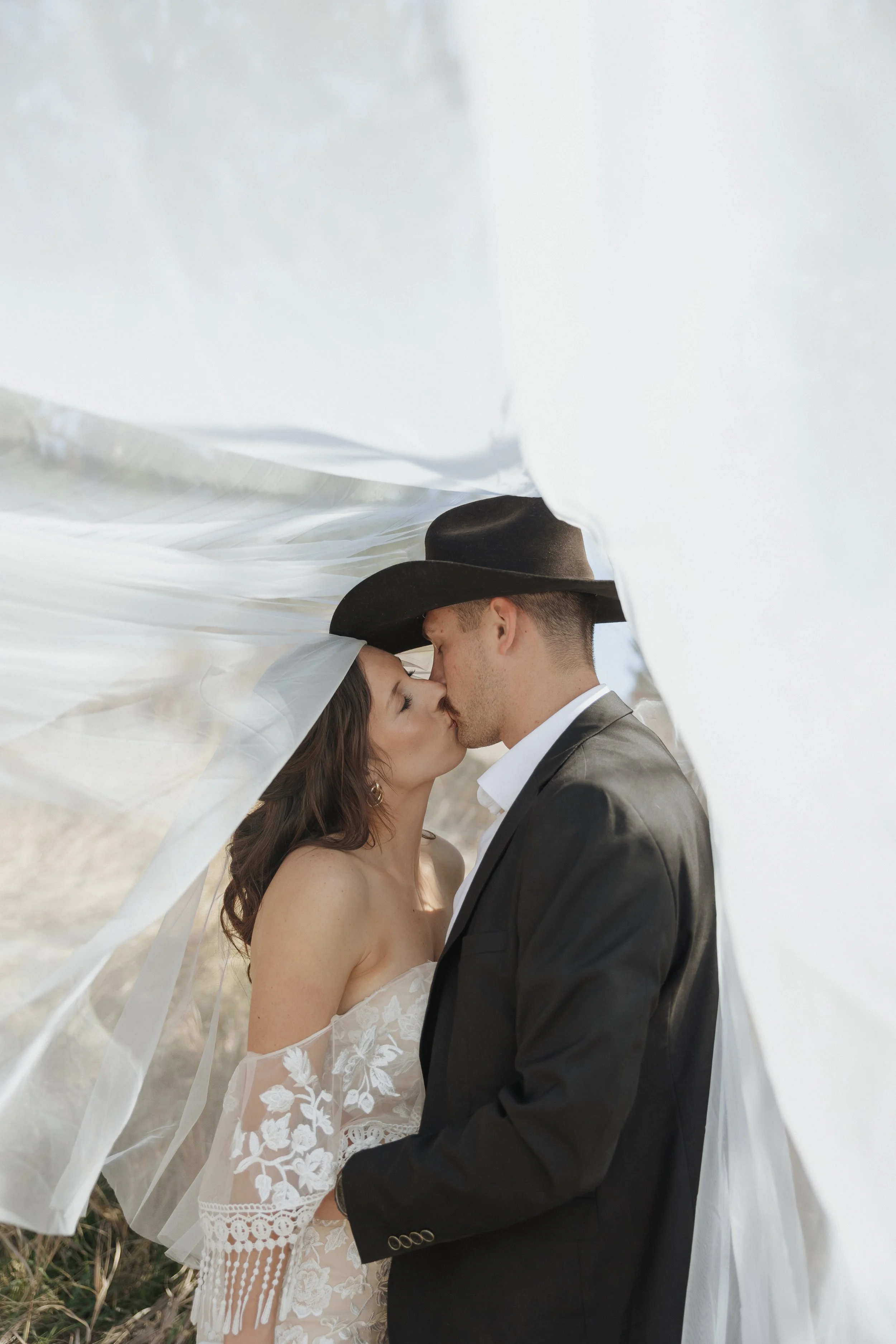 A bride and groom are kissing outdoors under a flowing white fabric canopy, with the bride wearing a lace wedding dress and the groom in a black suit and cowboy hat.