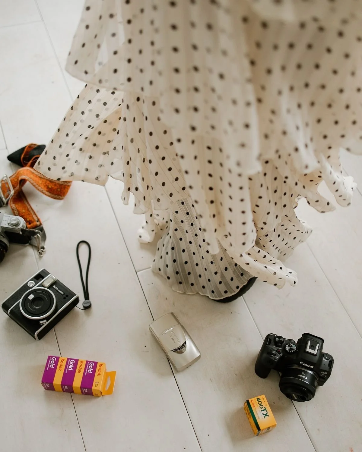 Various cameras, film rolls, and accessories scattered on the floor next to a polka dot dress hangin'}
