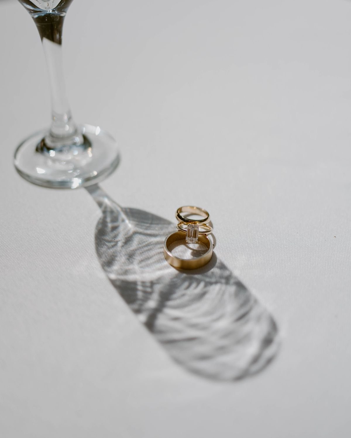 A close-up of two gold rings, one with a large rectangular gemstone, placed on a white surface. A wine glass is in the background, casting a shadow with reflections of the rings.