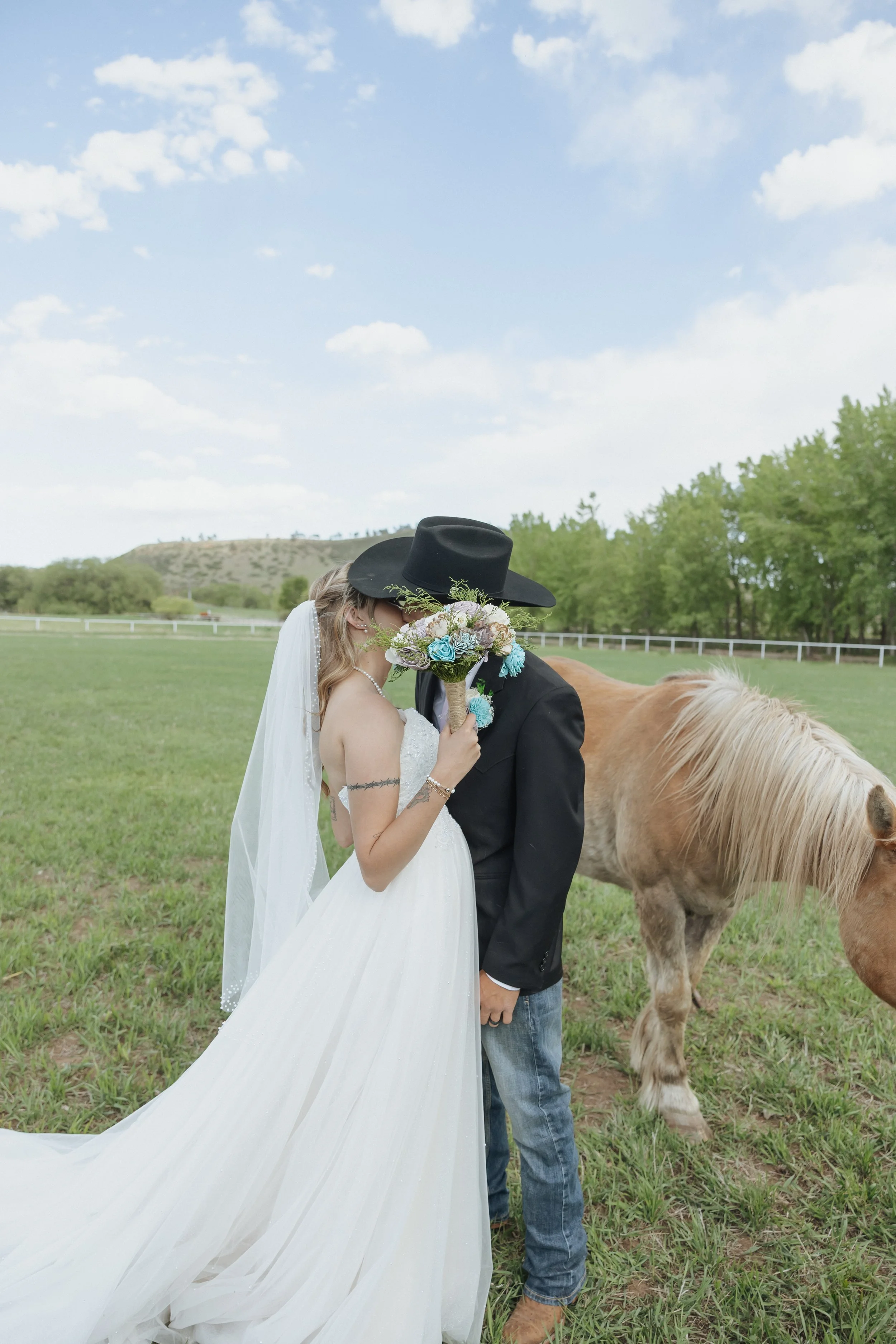 Couple kissing outdoors on a grassy field, woman in a white wedding dress and veil holding a bouquet, man in a black hat and black jacket, pony nearby, trees and blue sky in the background.
