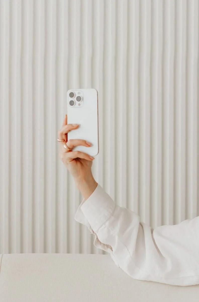 A person wearing a white long-sleeve shirt taking a mirror selfie with an iPhone in front of a textured beige wall.