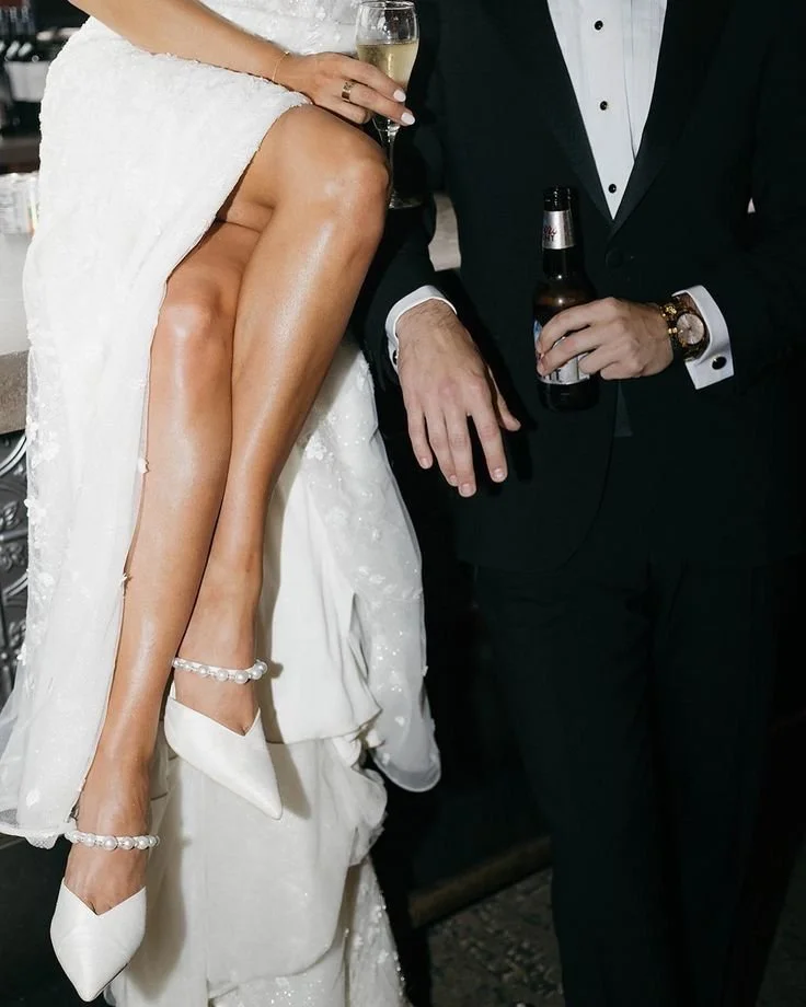 Close-up of a bride in a white gown and pearl-embellished shoes seated on a man's lap, holding a glass of champagne, with the man dressed in a tuxedo holding a beer bottle.