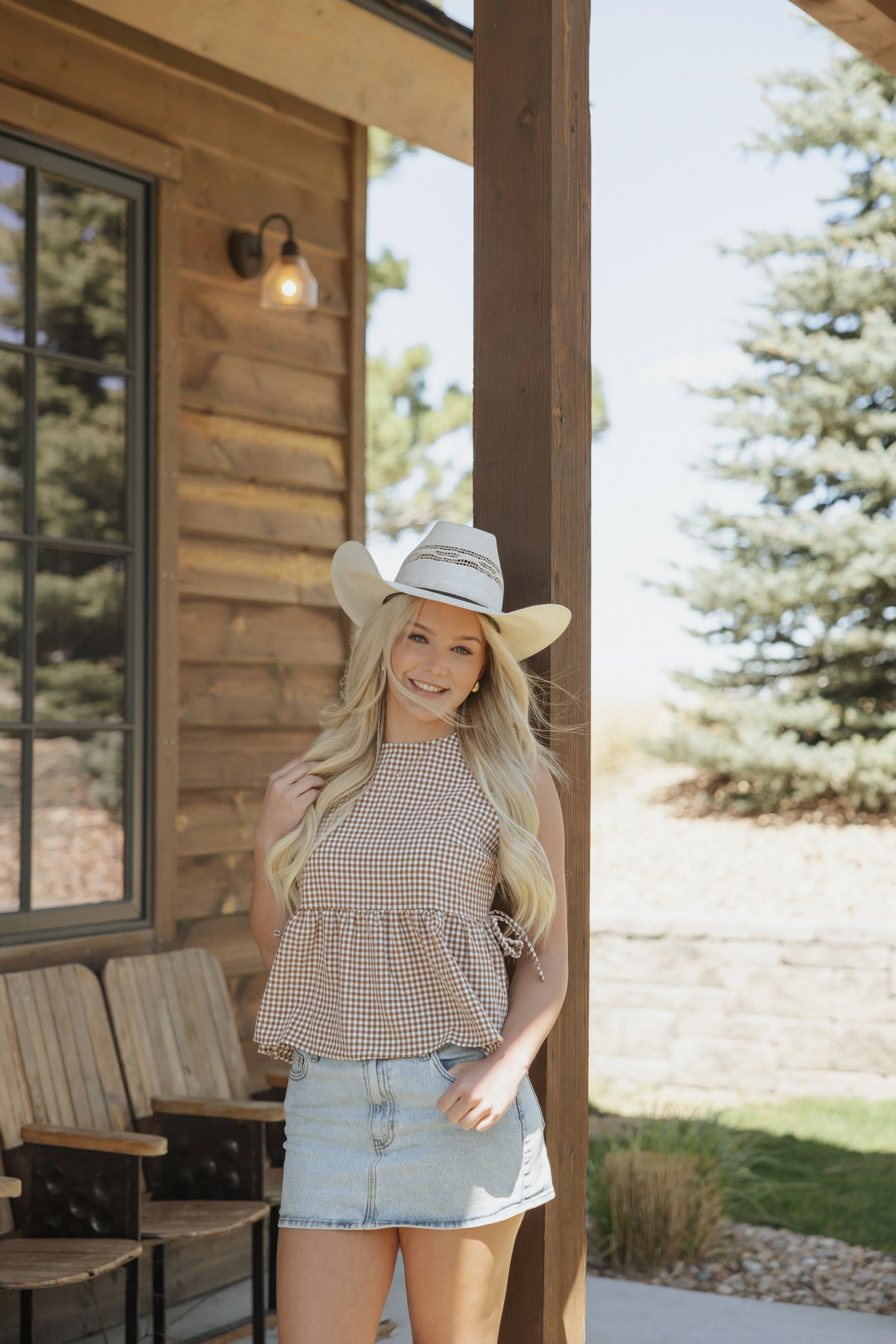 A young woman with long blonde hair wearing a wide-brimmed hat, a gingham sleeveless top, and a denim skirt standing outside a rustic wooden building with benches and trees in the background.