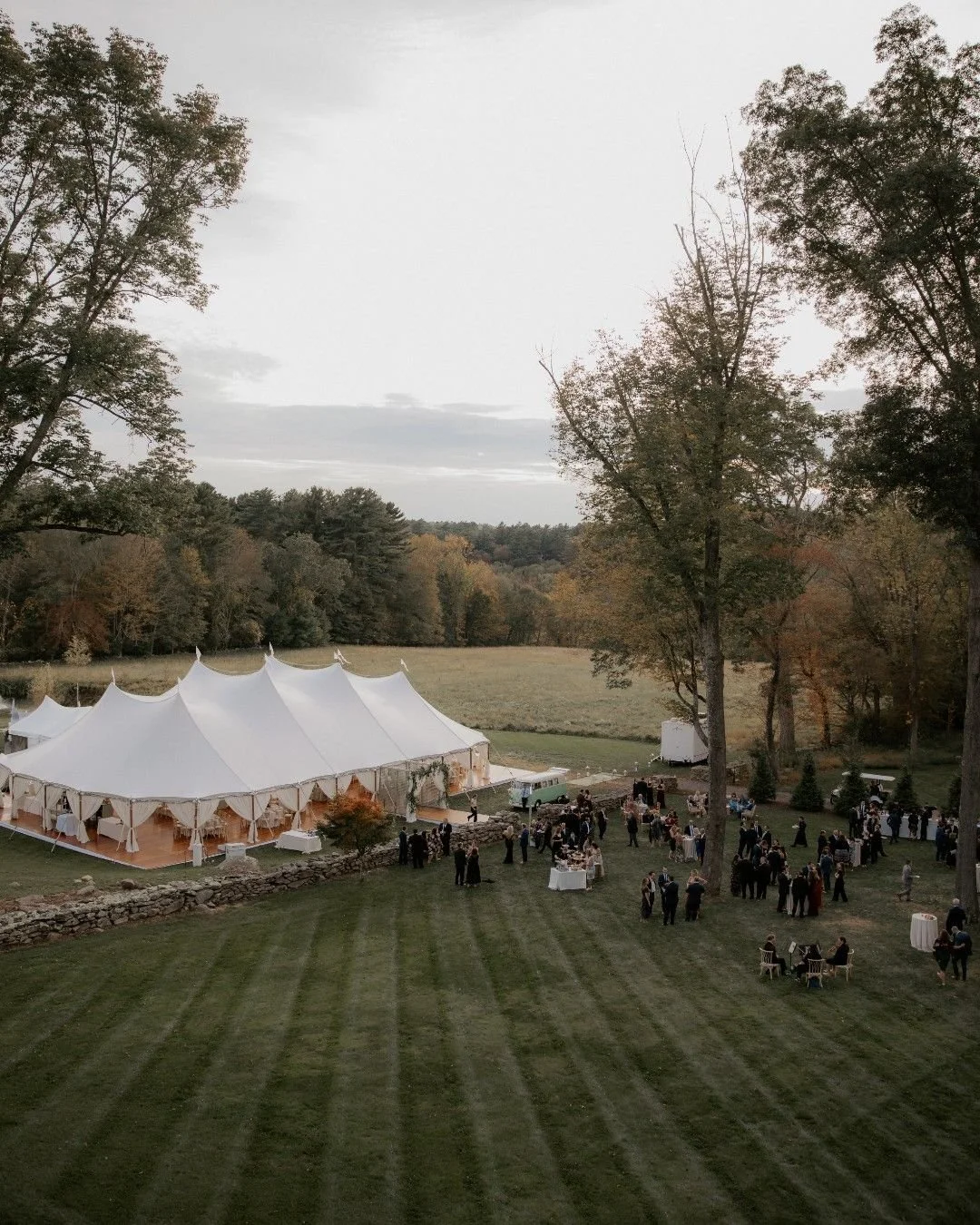Outdoor wedding reception with large white tent, tables, and guests on a lawn bordered by trees and a stone wall in the evening.