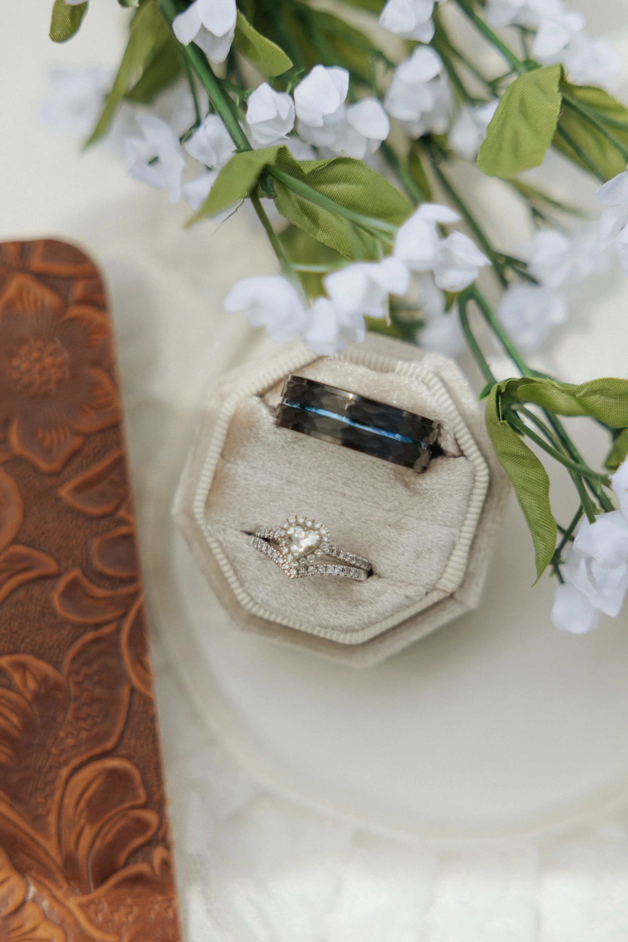 Close-up of a jewelry box with rings, placed on a white surface, with white flowers in the background and part of a brown bag visible on the side.