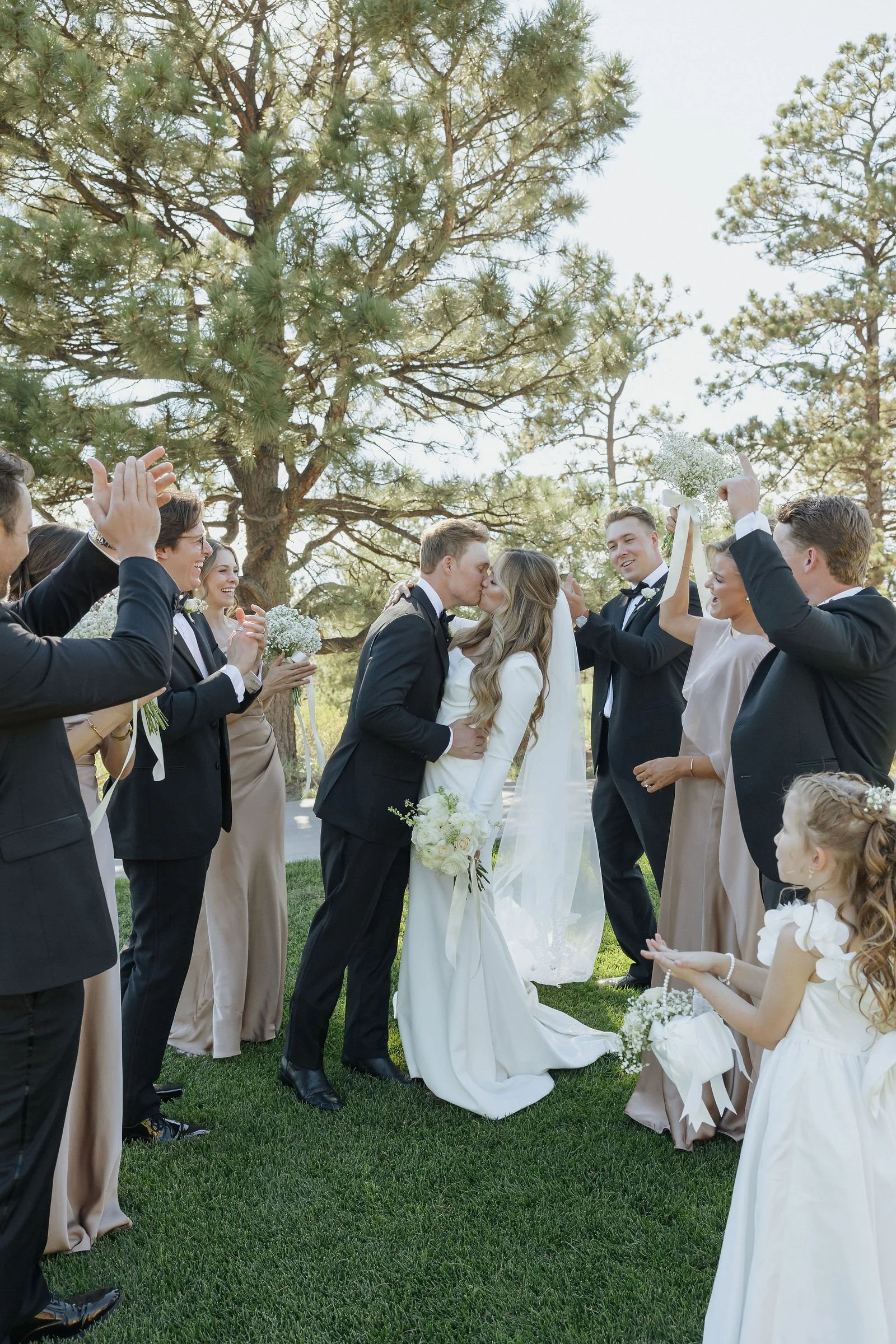 A wedding ceremony taking place outdoors under a large pine tree, with a bride and groom kissing surrounded by friends and family.