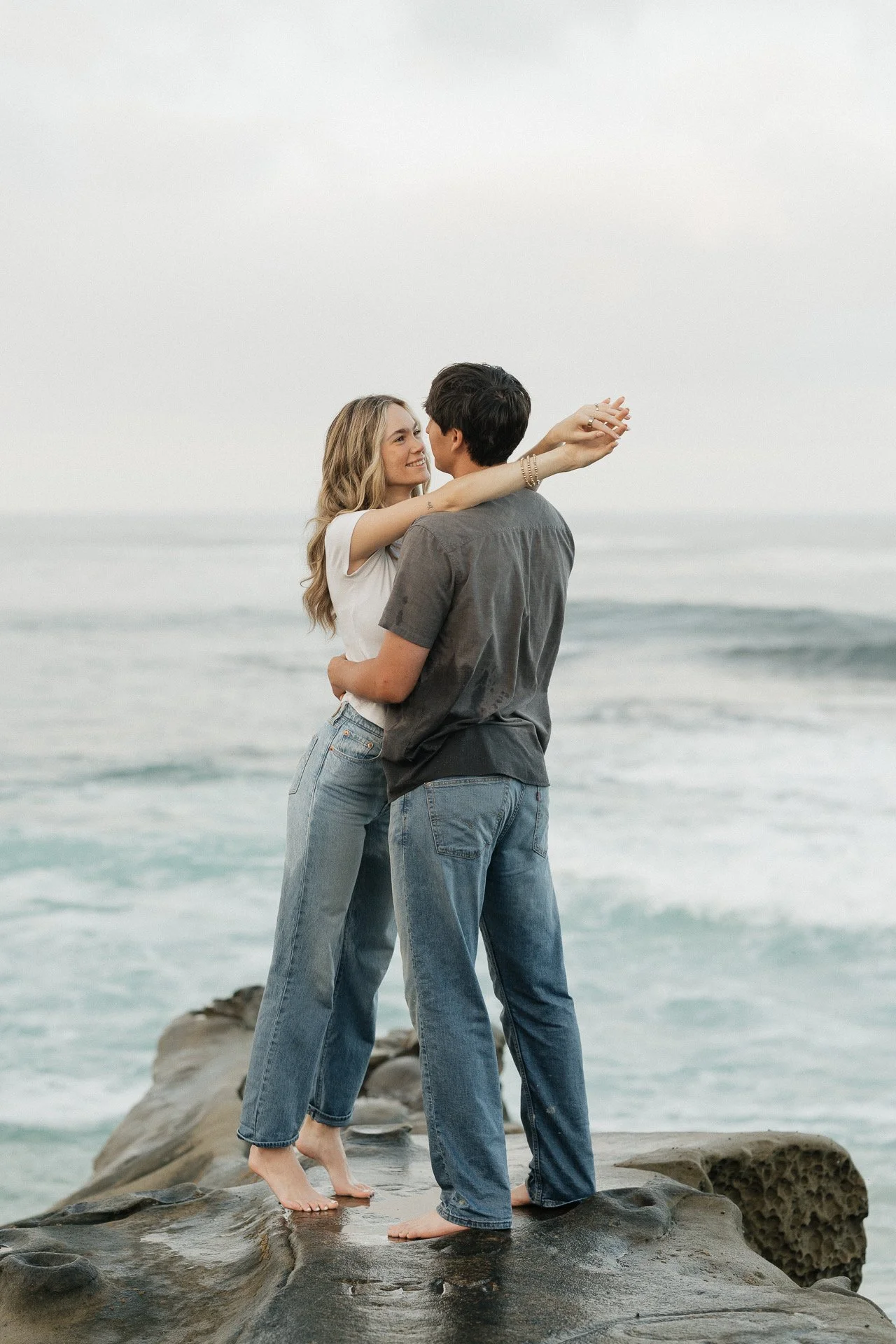 A couple standing on rocks at the beach, embracing and looking into each other's eyes, with the ocean in the background.