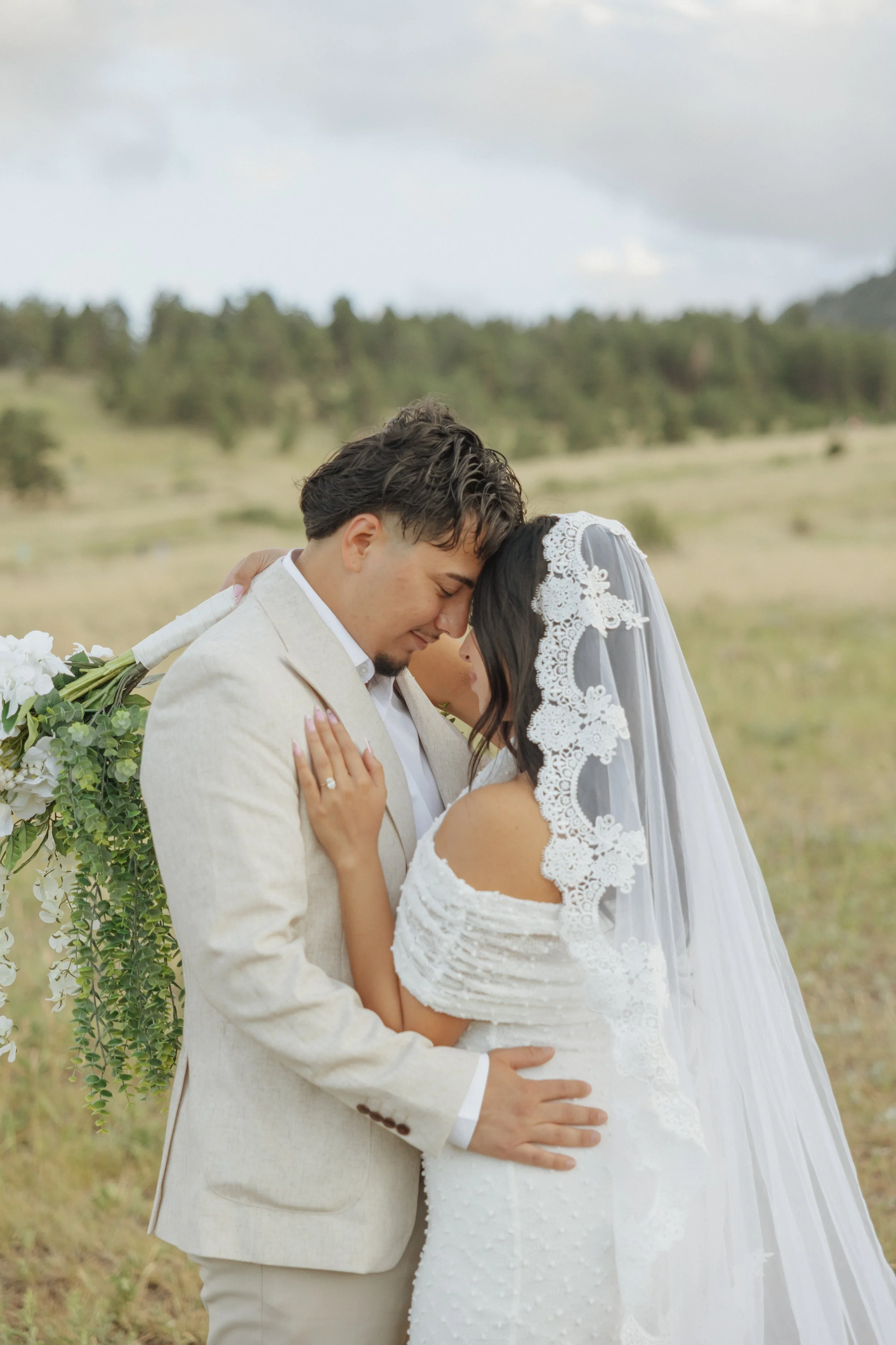 A newlywed couple embraces in a field, with the groom wearing a beige suit and the bride in a lace wedding dress with a veil, standing close with their foreheads touching.