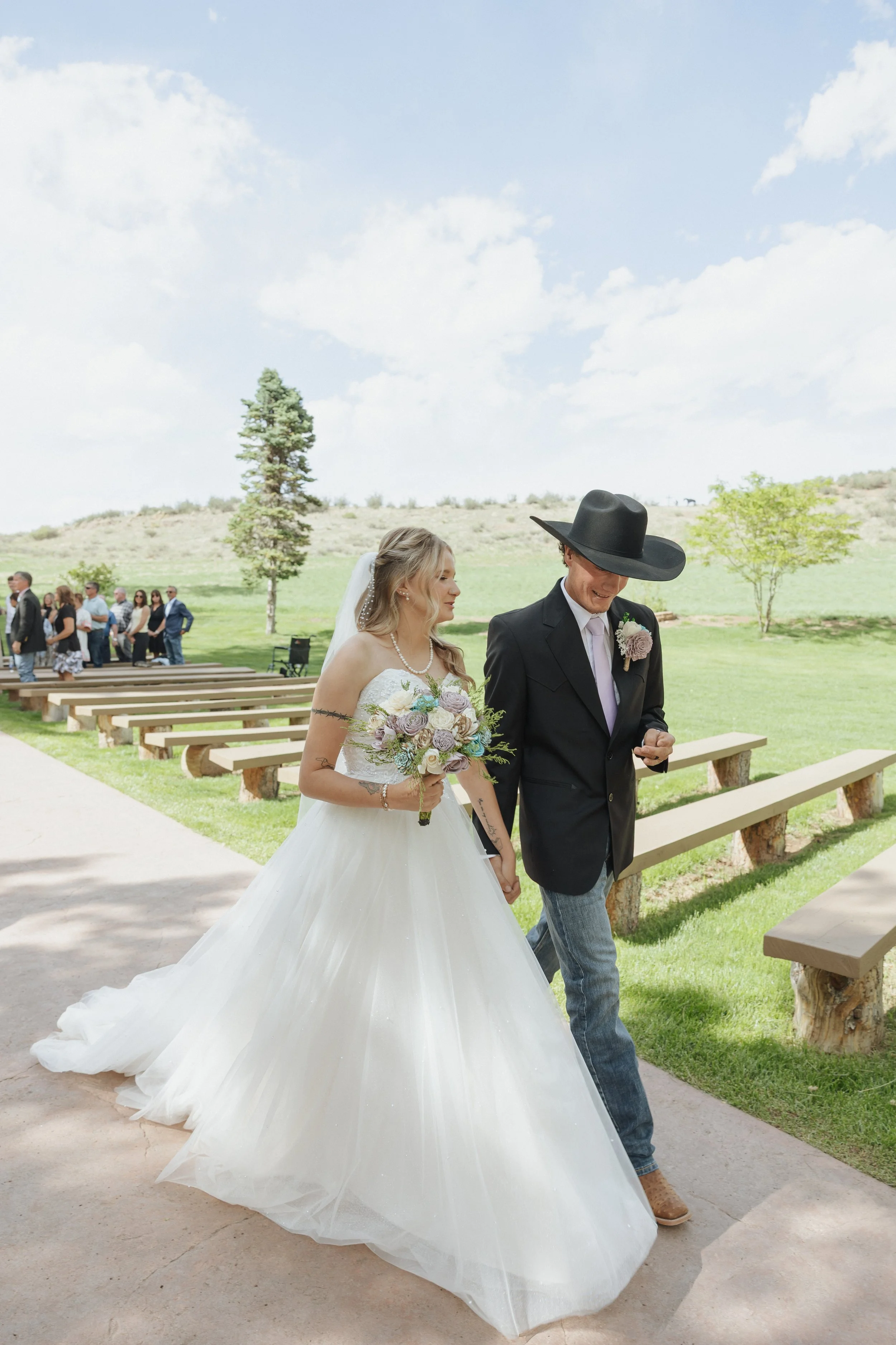 A bride and groom walking outdoors during a wedding ceremony on a sunny day. The bride wears a white wedding gown and holds a bouquet of flowers, while the groom wears a black suit with a wide-brimmed hat and a boutonniere. Guests are seated and stan