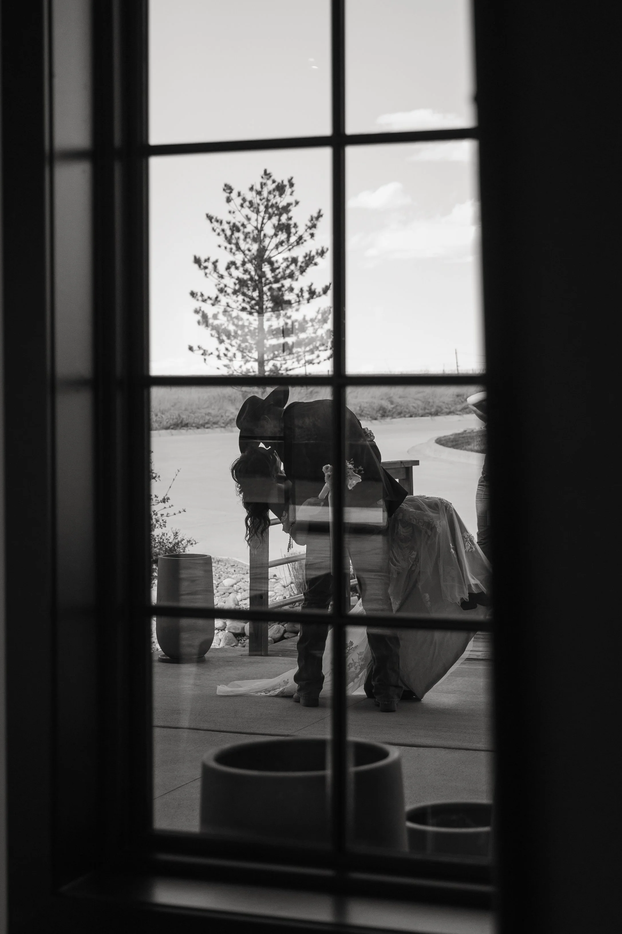 View through a window showing a wedding ceremony outdoors with a bride in a wedding dress bending over and a man in a suit. A tree and a cloudy sky are visible in the background.