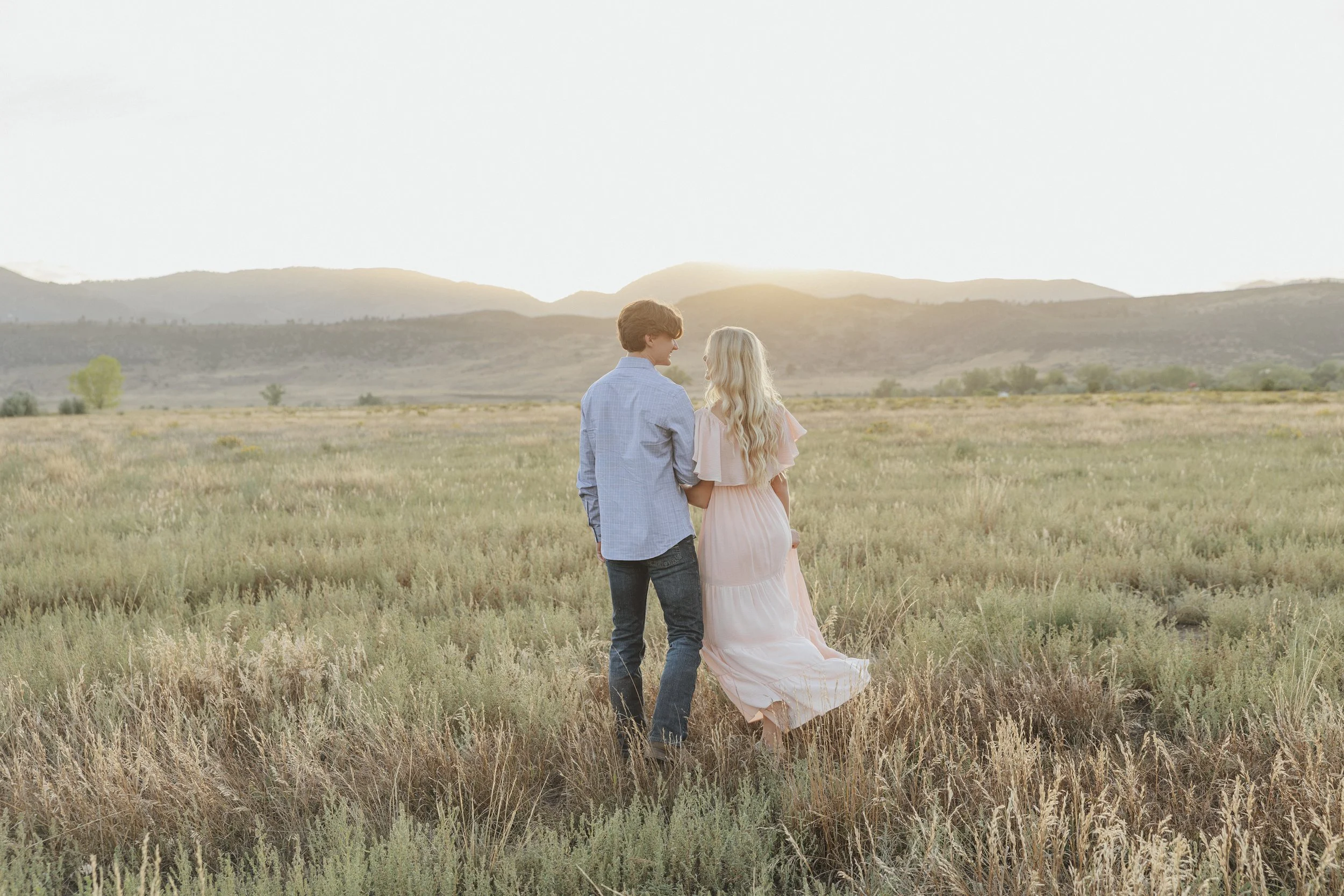 A couple holding hands and looking at each other in a grassy field with mountains in the background during sunset.