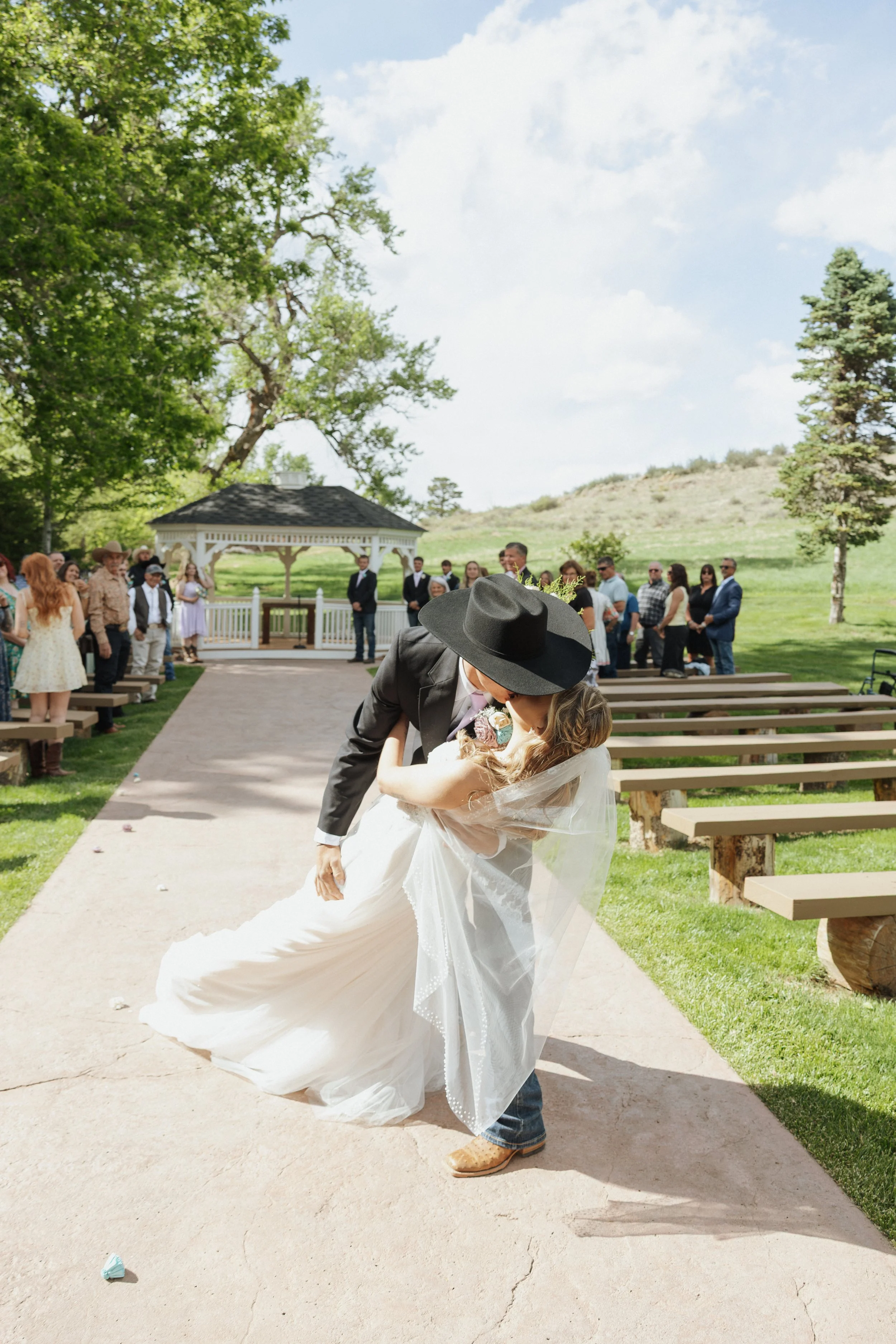 A bride and groom sharing a kiss during an outdoor wedding ceremony at a park. The bride wears a white wedding dress with a veil, and the groom wears jeans, a jacket, and a black hat. Guests are seated and standing in the background, with trees and a