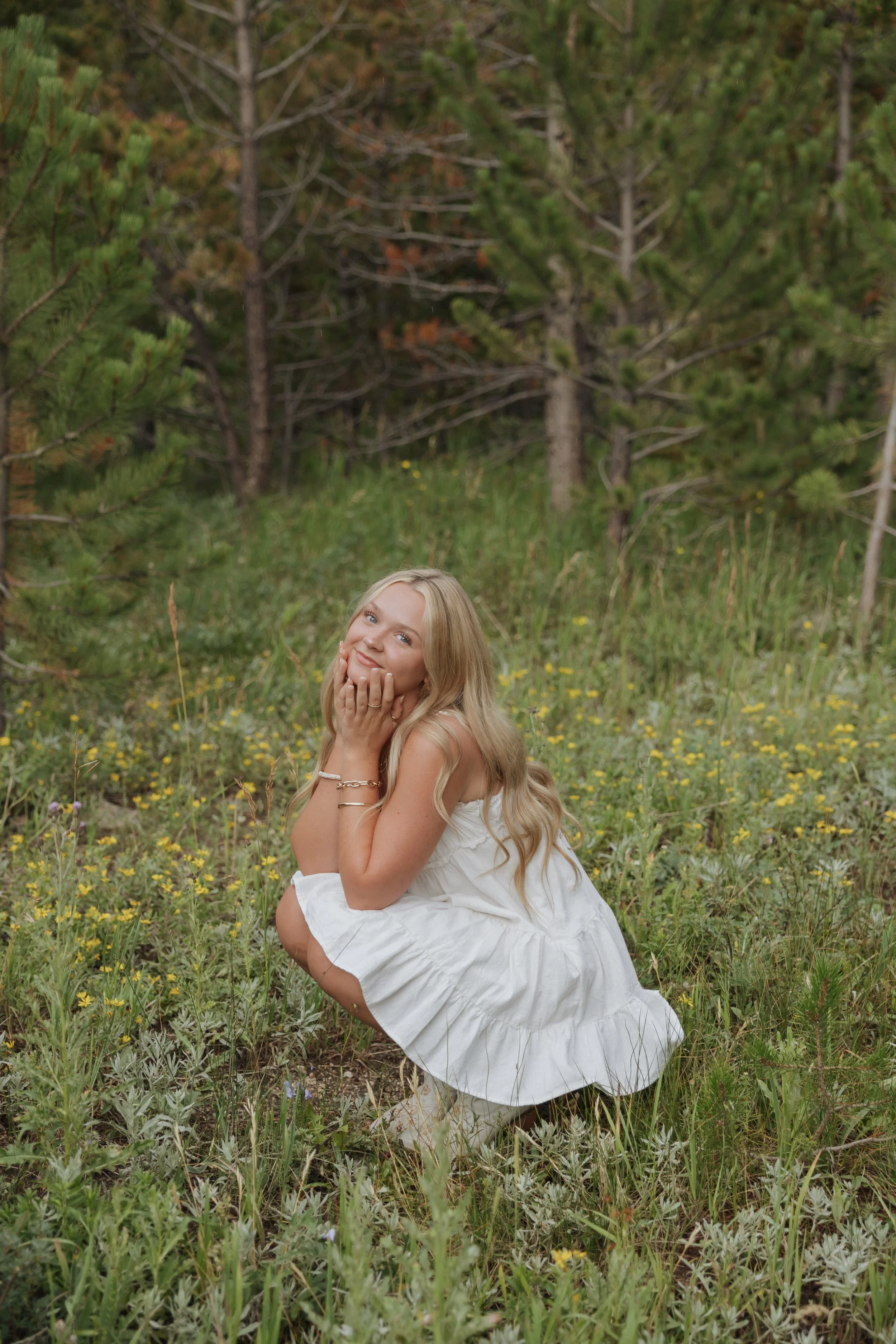 Young woman in a white dress crouching in a grassy field with yellow flowers, surrounded by trees.