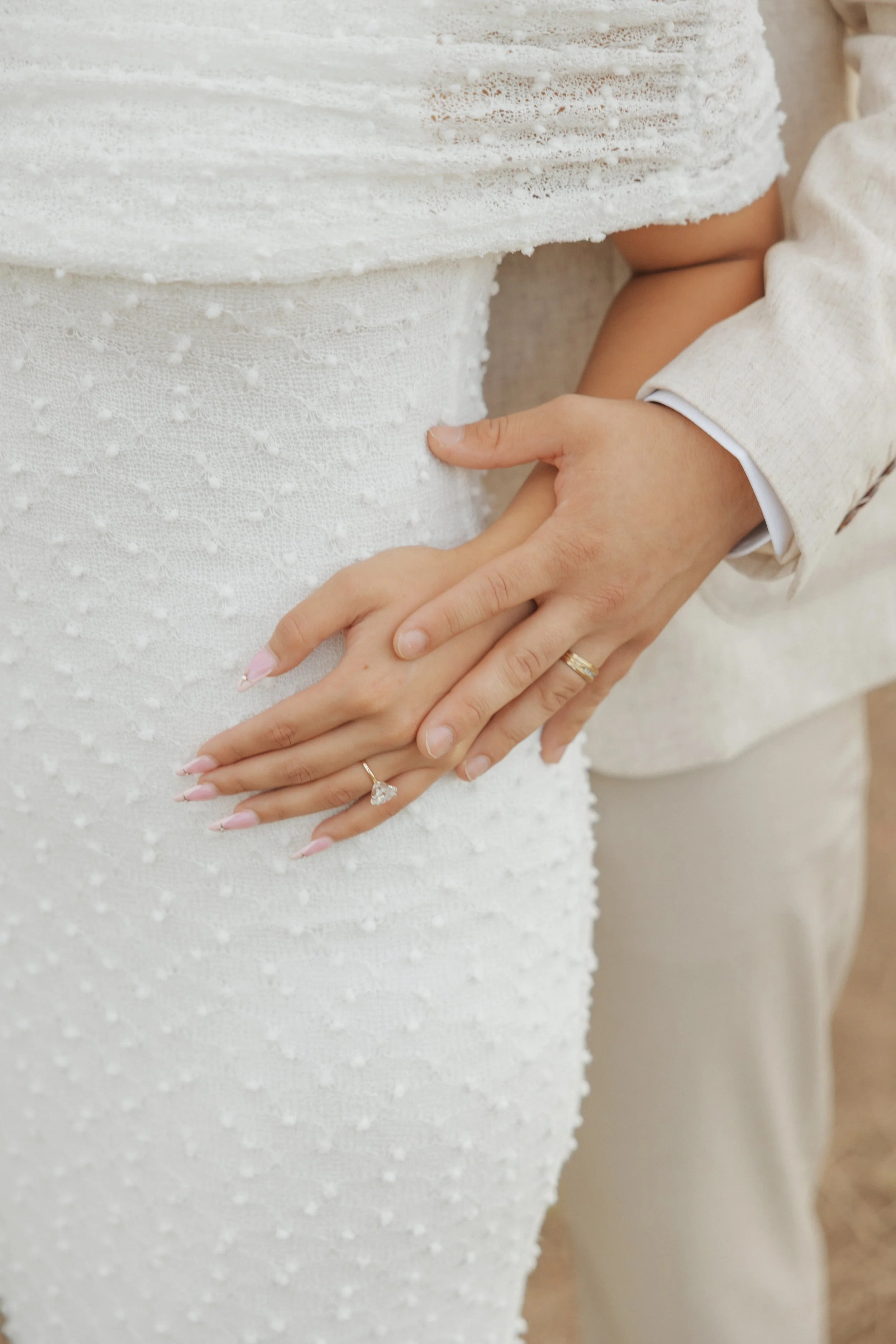 Close-up of a woman's hand with a wedding ring, gently resting on a bride's waist, dressed in white with textured lace fabric. A man in a light-colored jacket touches her waist with his hand.
