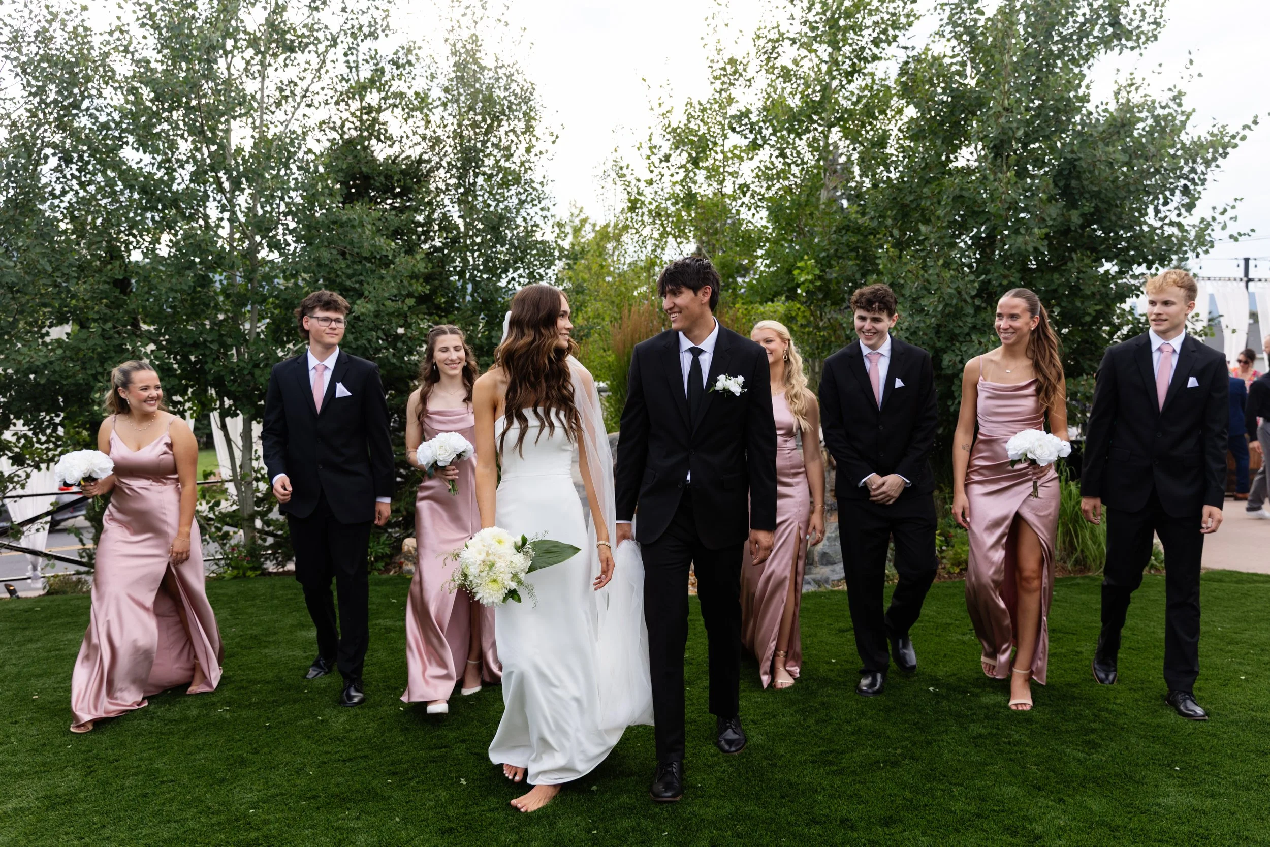 Group of people walking outdoors at a wedding, with greenery and trees in the background.