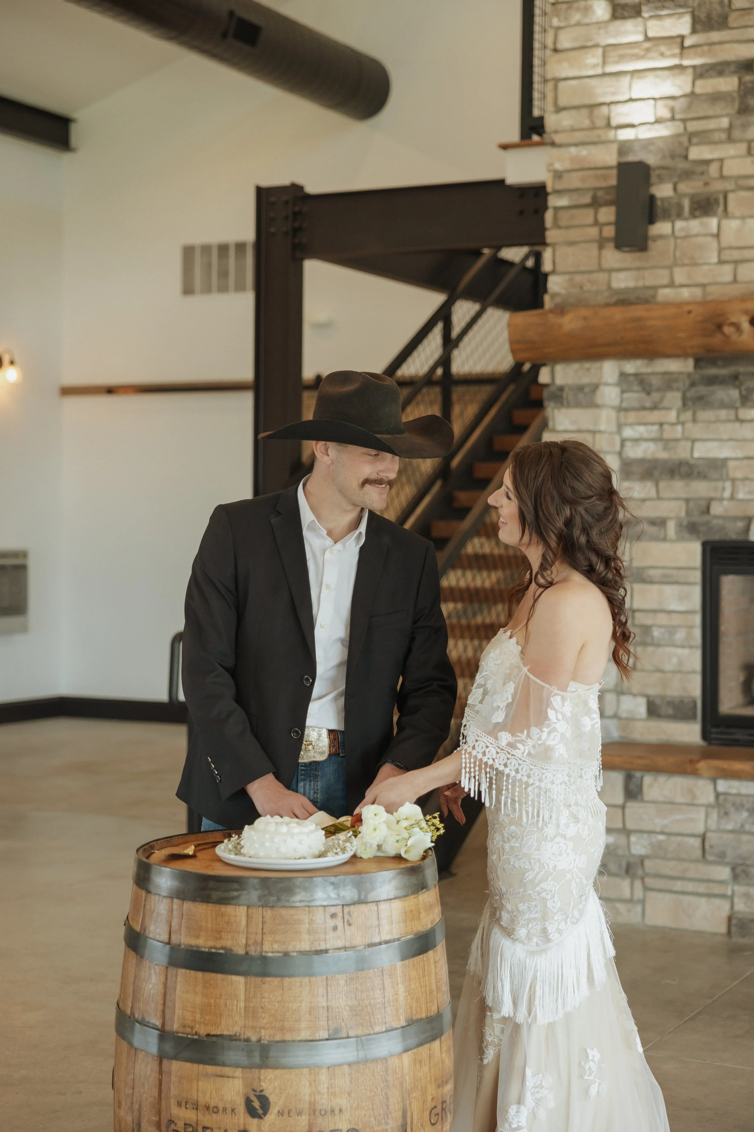 A groom and bride sharing a moment during a wedding ceremony indoors. The groom is wearing a black suit and a cowboy hat, while the bride is in a white lace wedding dress. They are standing behind a wooden barrel with a cake on top, and holding hands