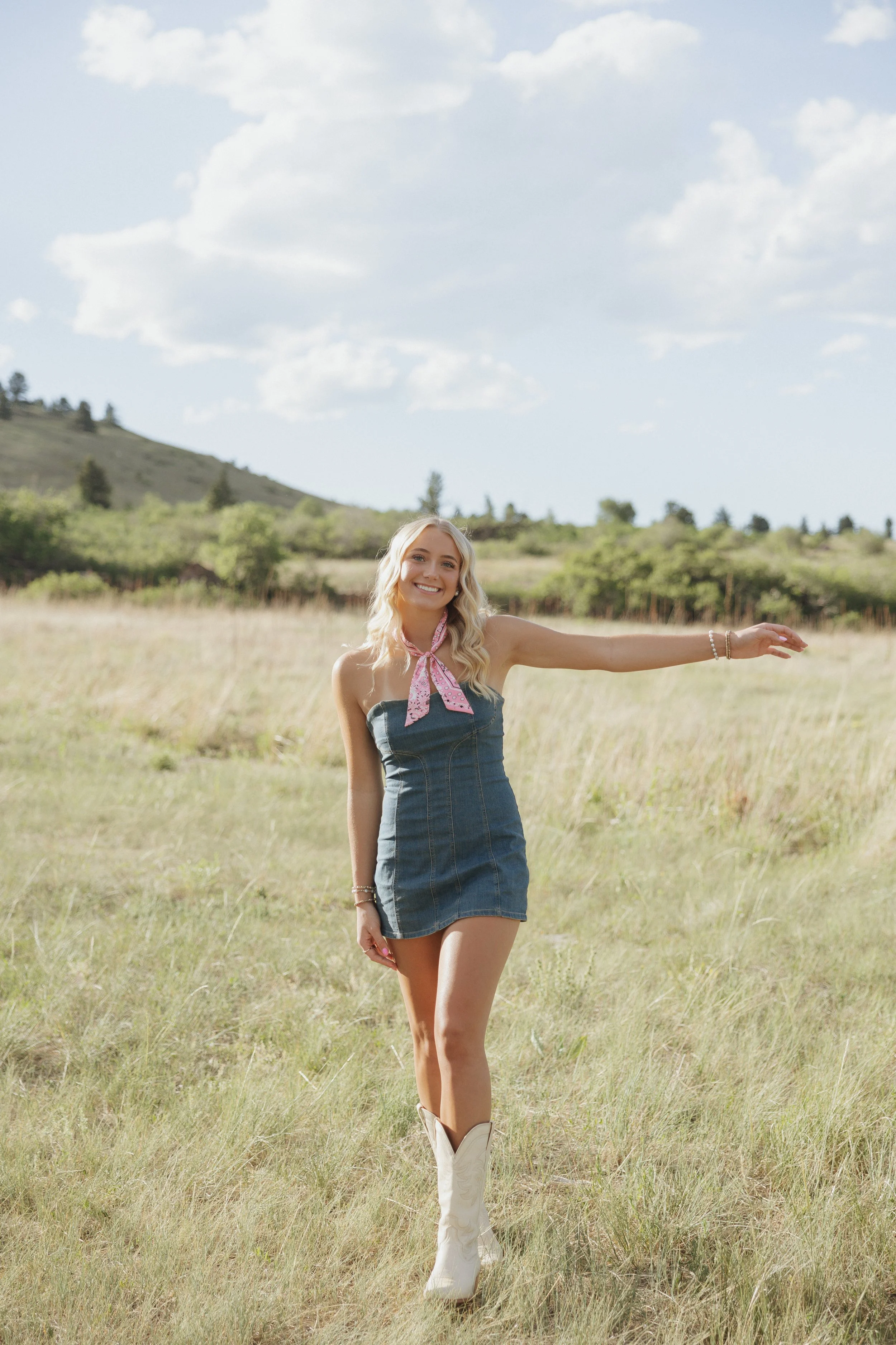A young woman with long blonde hair smiling in a field on a sunny day, wearing a denim dress, white cowboy boots, and a pink neck scarf.