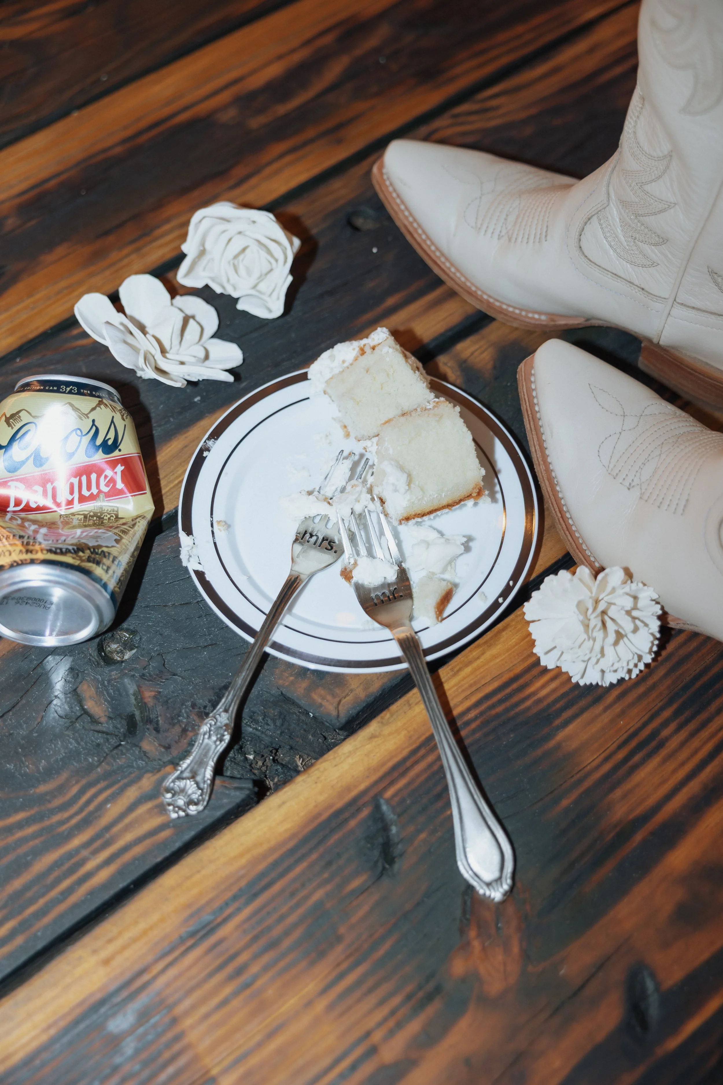 Close-up of cowboy boots, a partially eaten slice of cake on a plate, a soda can, and crumpled paper flowers on a rustic wooden table.