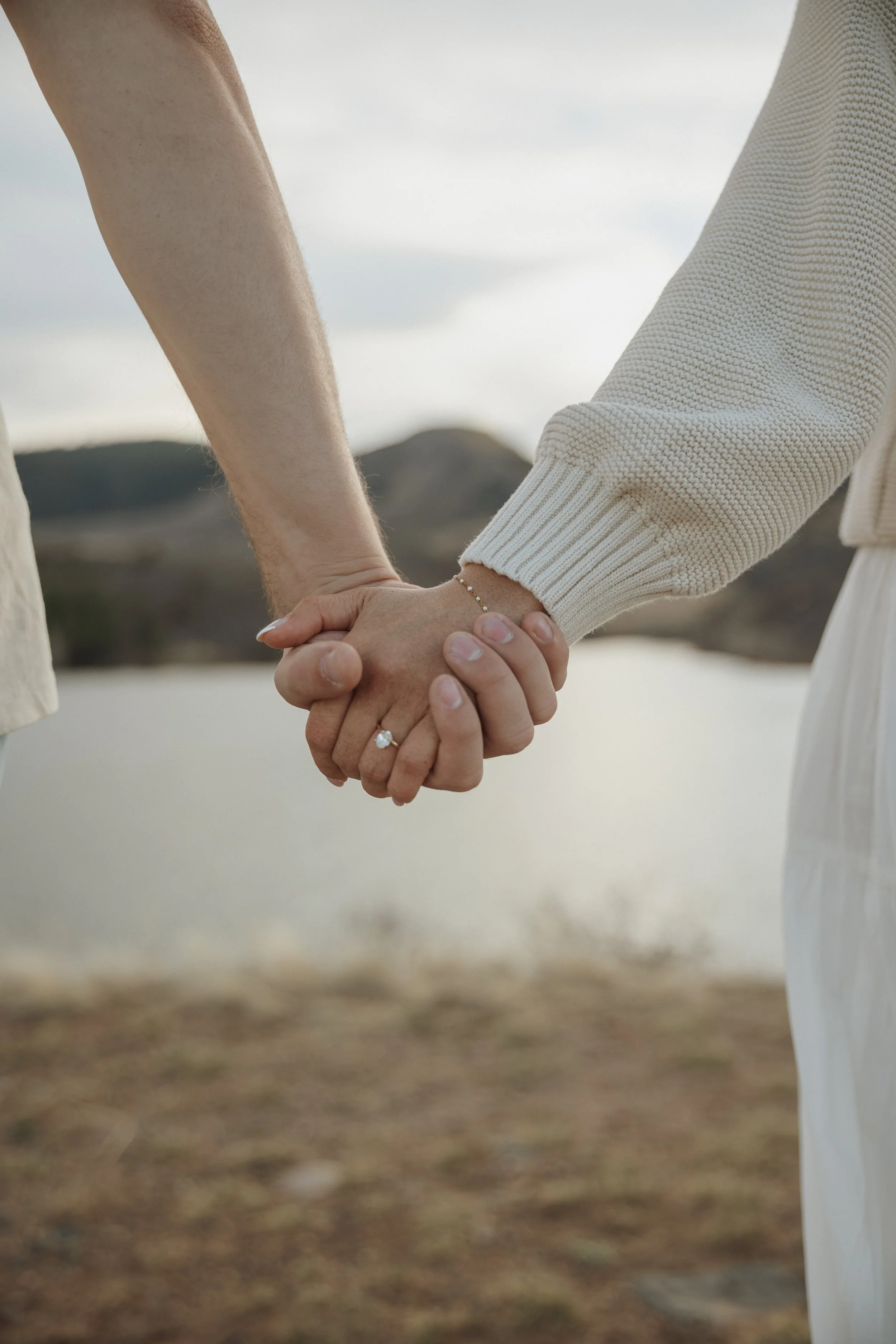 Close-up of two people holding hands outdoors near a body of water with a mountainous landscape in the background, one wearing a ring and a sweater.