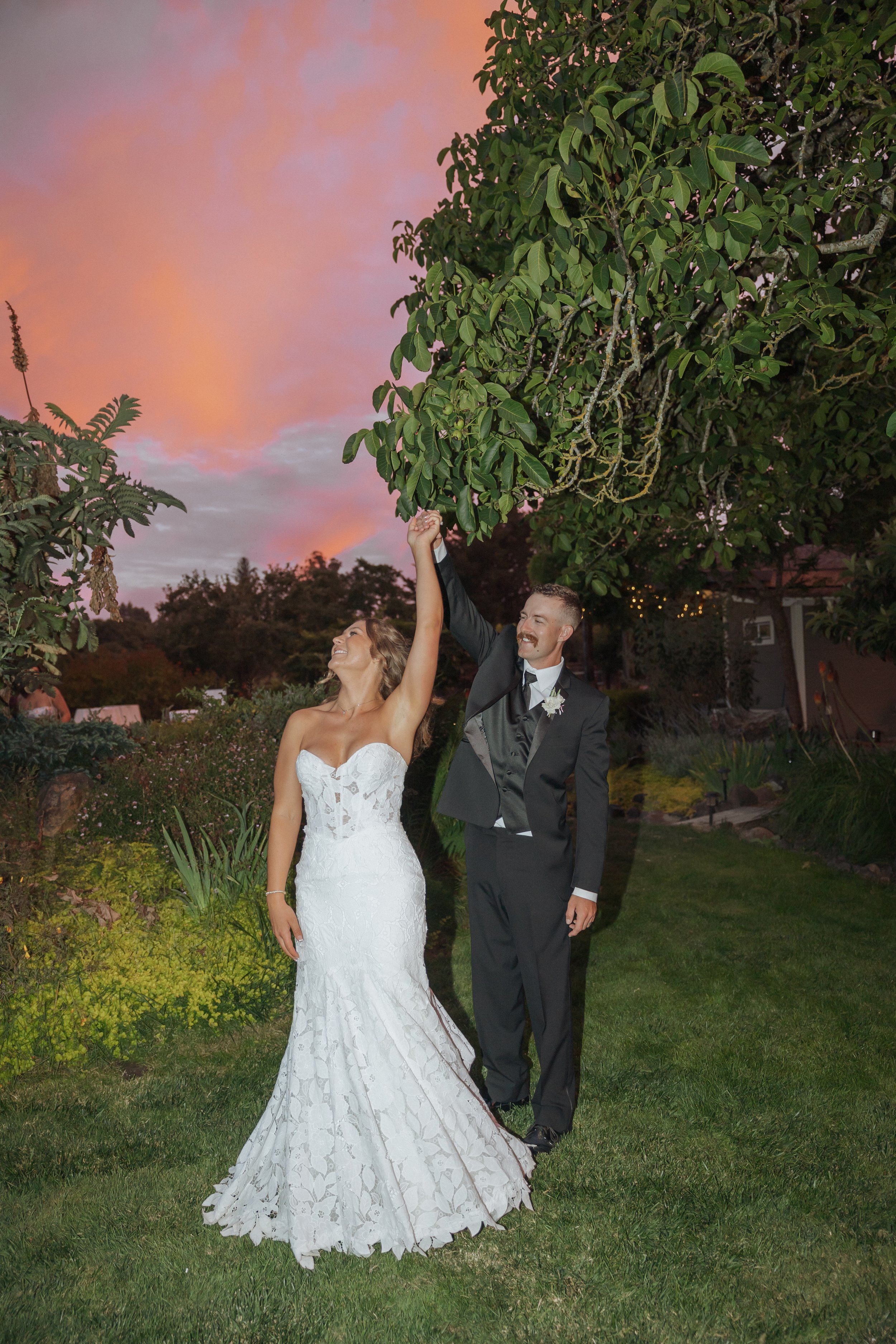 A bride and groom dancing outdoors in the evening with a pinkish sky in the background. The bride is wearing a white lace wedding gown, and the groom is in a black suit with a boutonniere. They are holding hands and smiling.