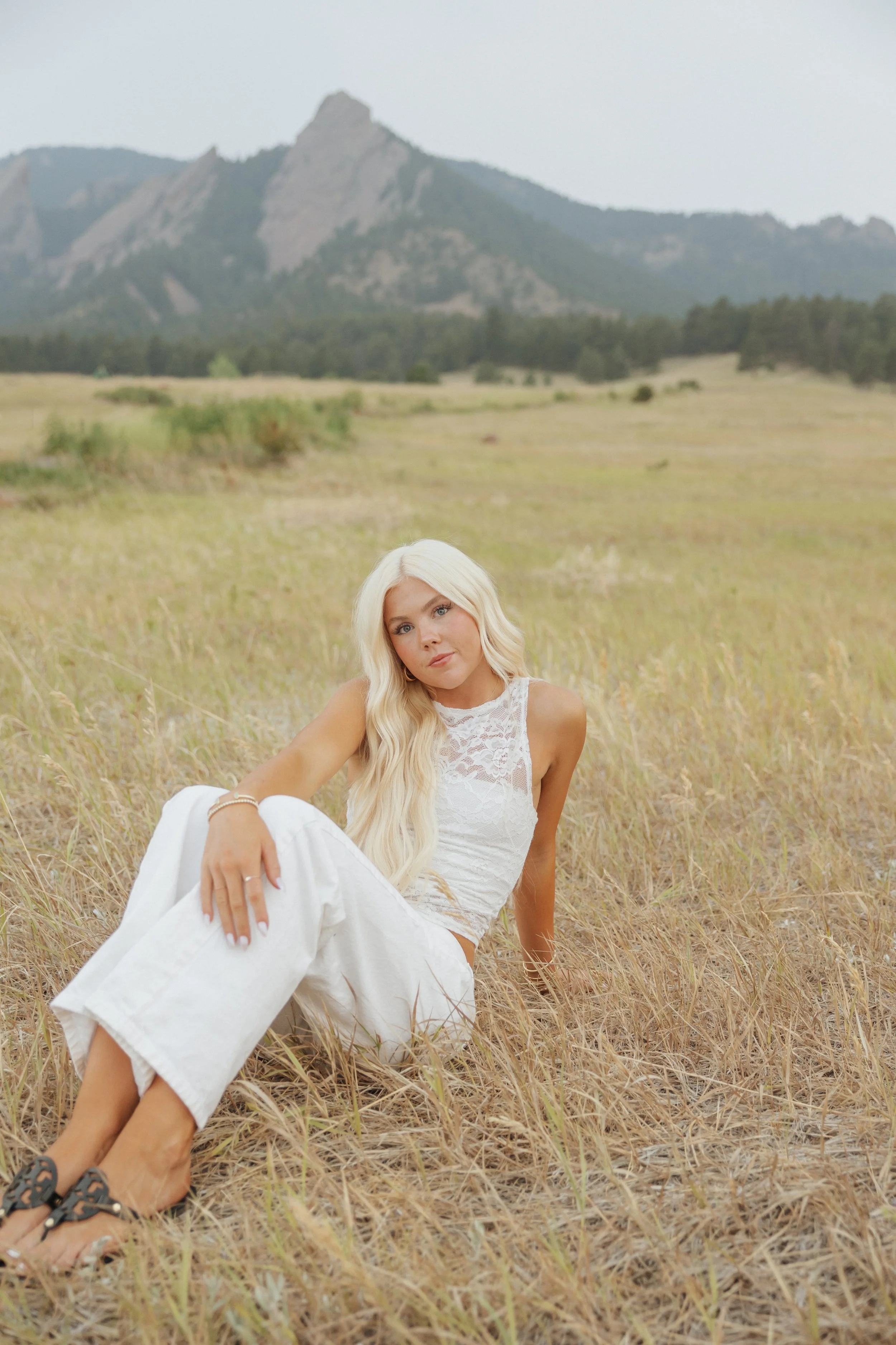A woman with long blonde hair sitting in a field of tall grass, with mountains in the background.