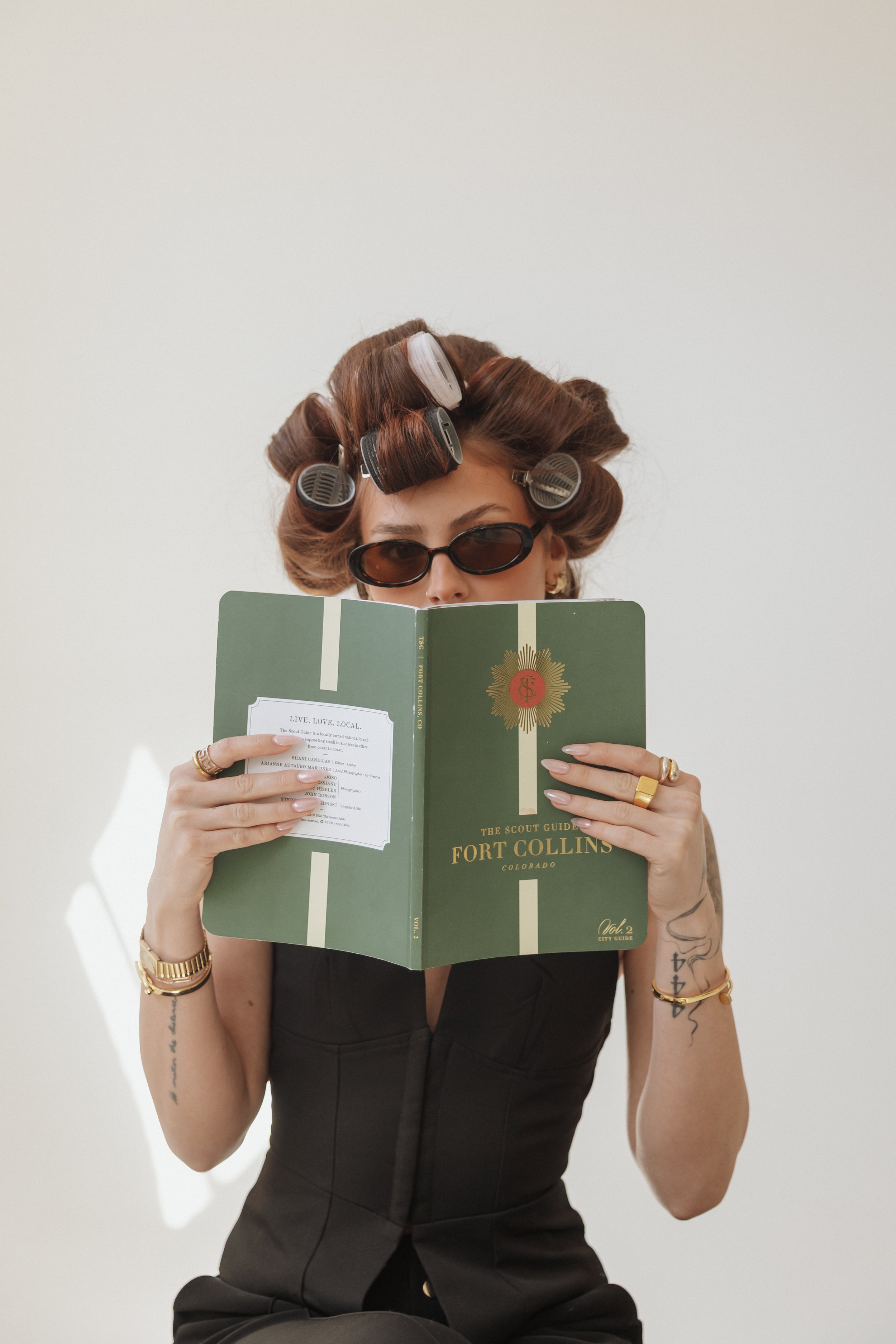 Woman with curlers in her hair and wearing sunglasses, reading a green book titled 'The Scout Guide Fort Collins, Colorado'.