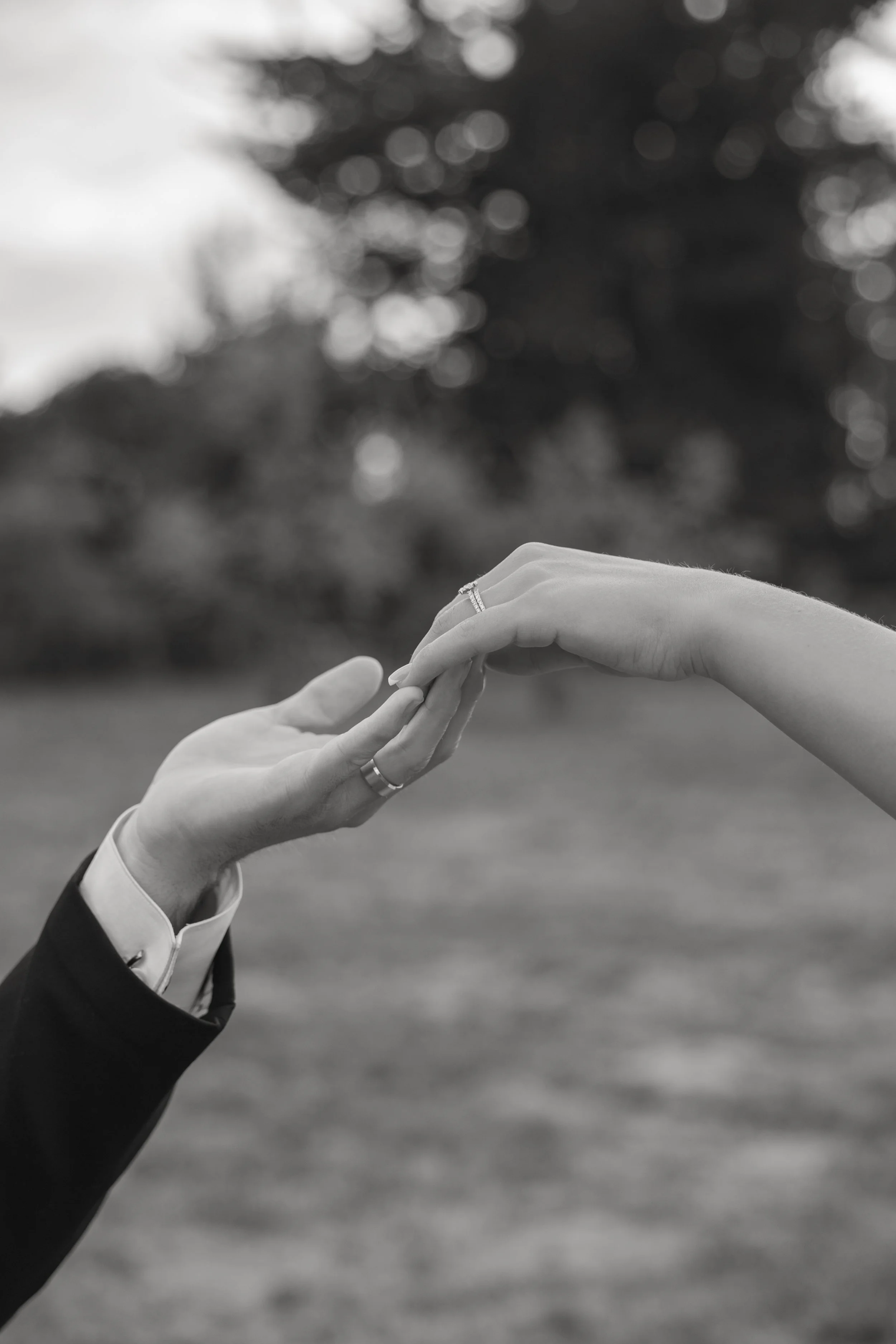 A close-up black and white photo of a groom in a suit with a boutonniere, holding hands with the bride, who is wearing a dress and a ring, outdoors with blurred trees in the background.