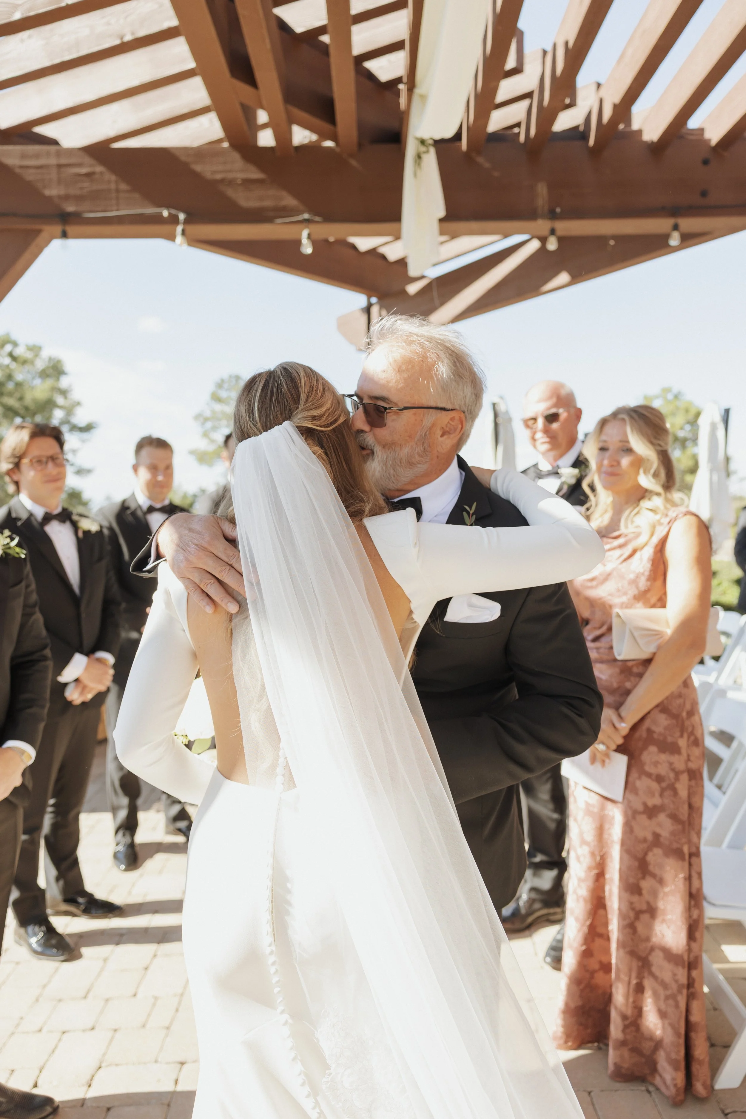 A bride and an older man, possibly her father, sharing a kiss at her wedding, surrounded by wedding guests outdoors.