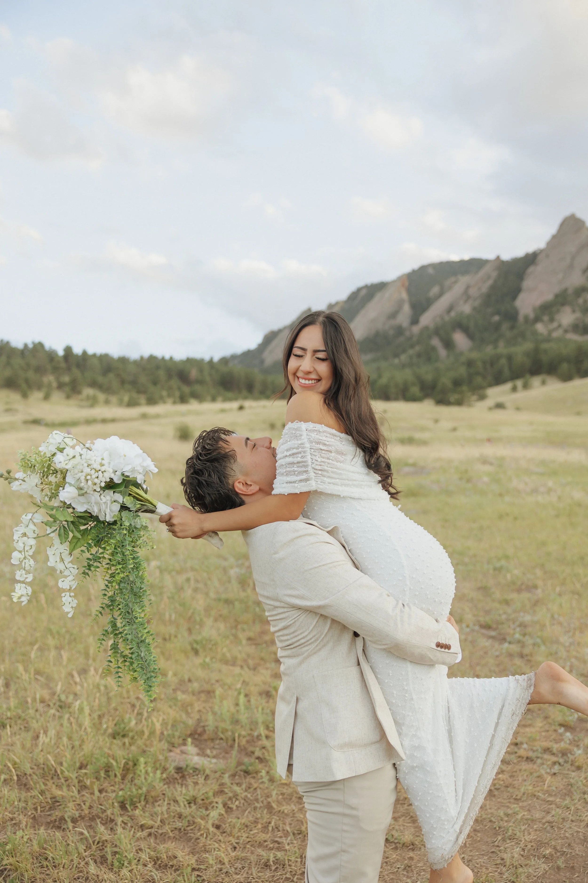 A joyful couple is outdoors in a field, with the man lifting the woman in his arms. The woman is smiling, wearing a white dress, and holding a bouquet of white flowers. The man is dressed in a light beige suit. There are mountains and trees in the ba