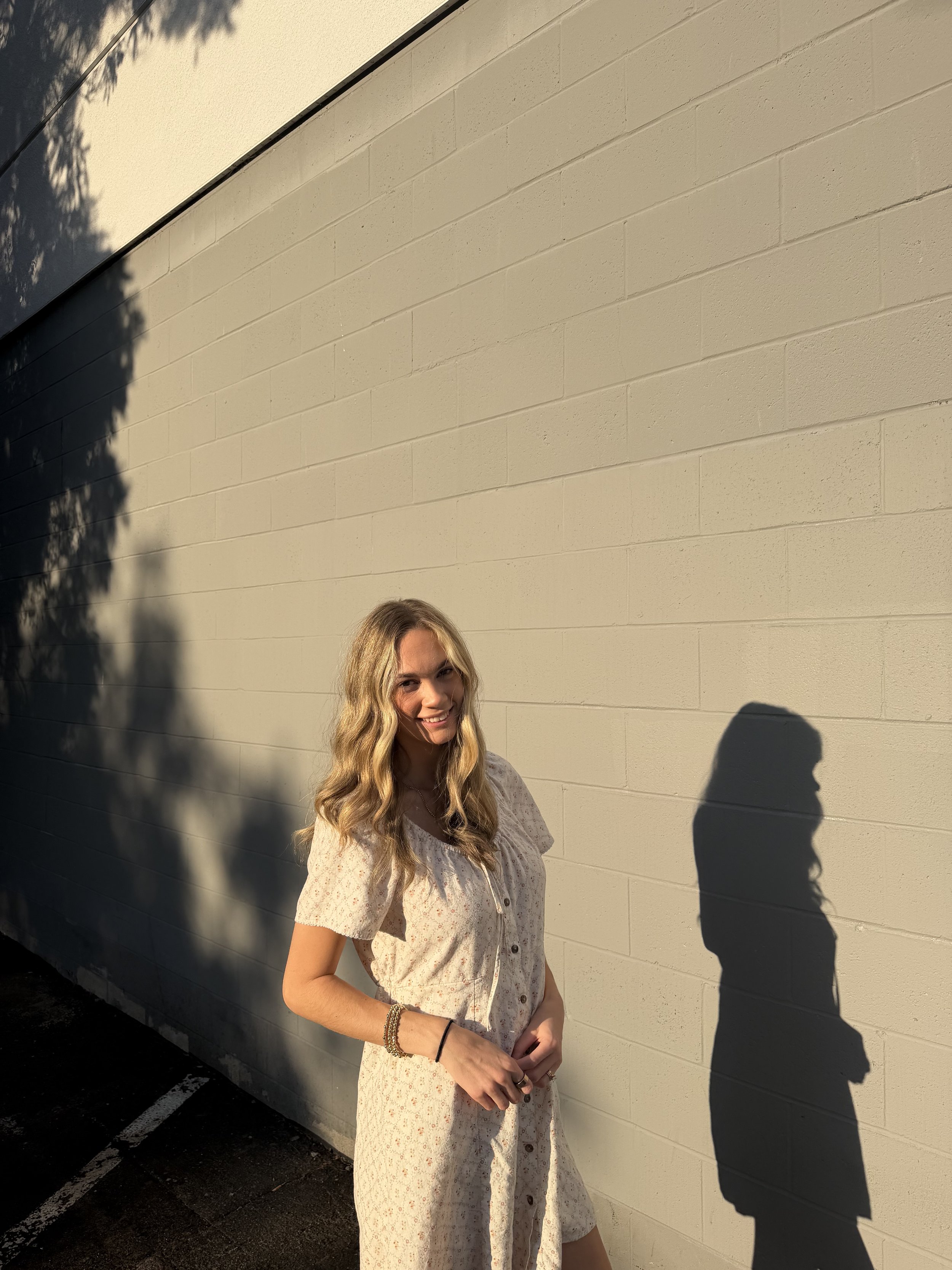 A woman with long wavy hair smiling, standing against a beige brick wall outside. The sunlight creates shadows on the wall, and her shadow is visible on the right side of the image.