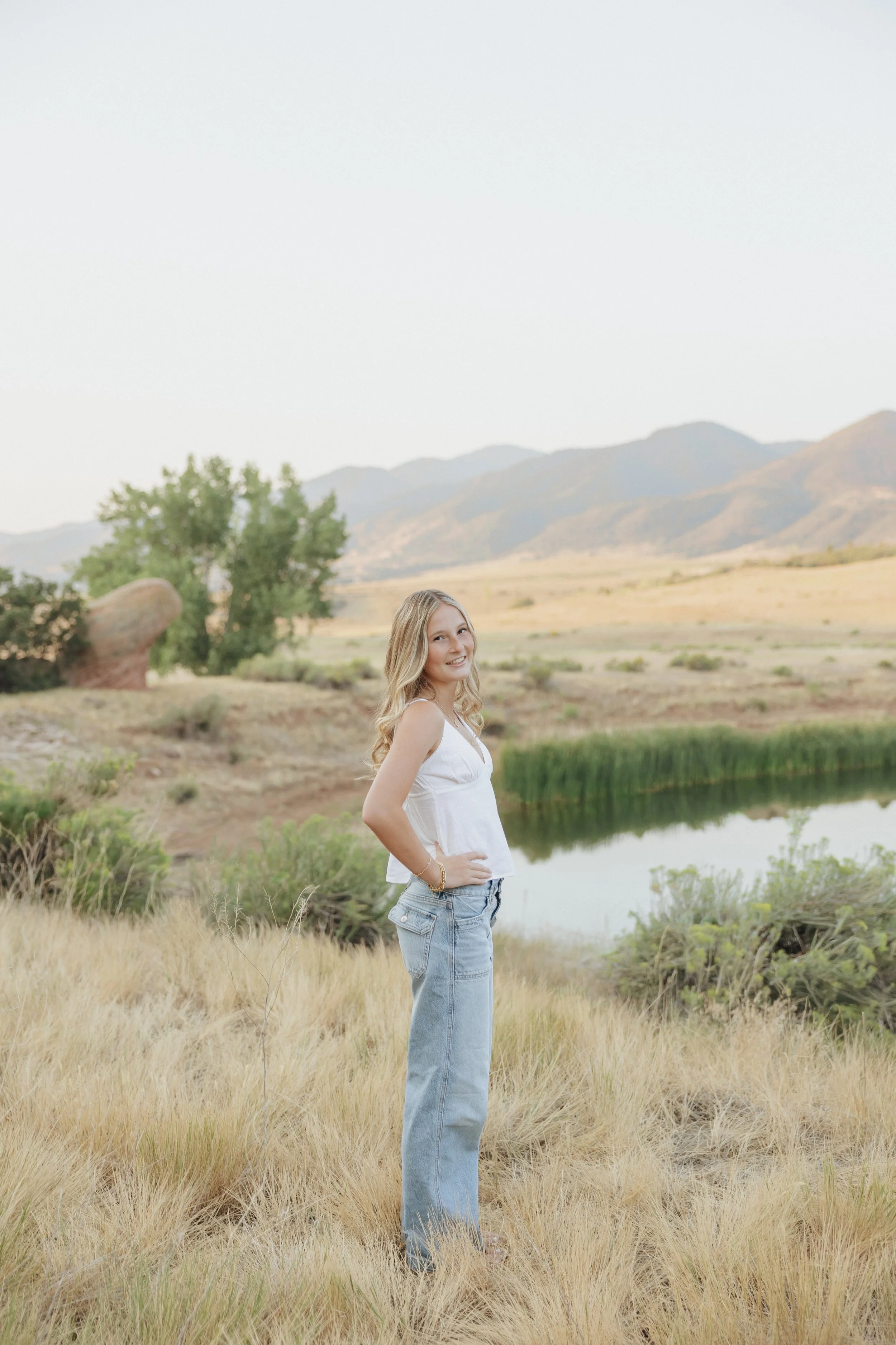 A young woman with blonde hair smiling, standing in a grassy field near a small body of water with mountains in the background.