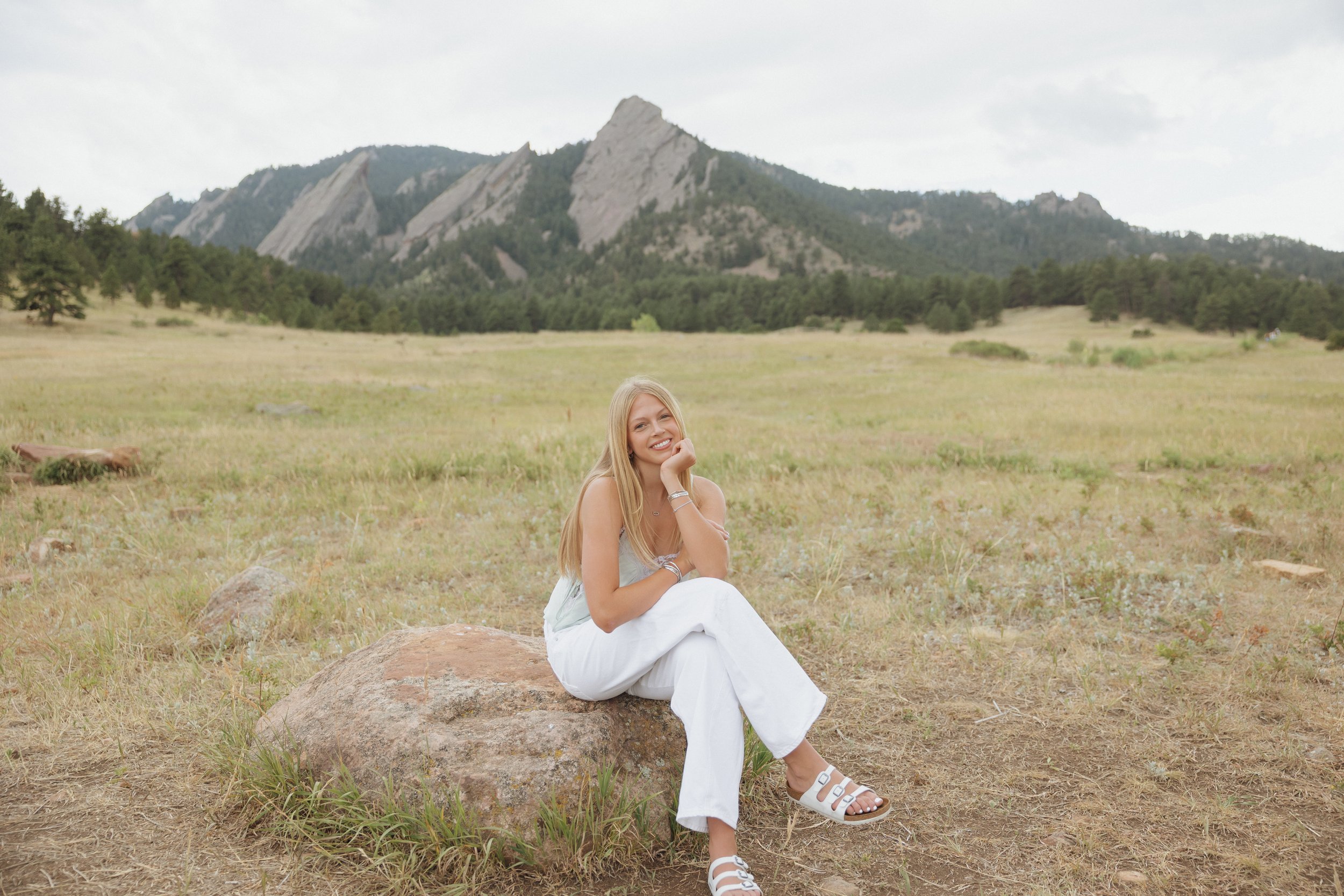 A young woman with long blonde hair sitting on a large rock in a grassy field with trees and mountains in the background.
