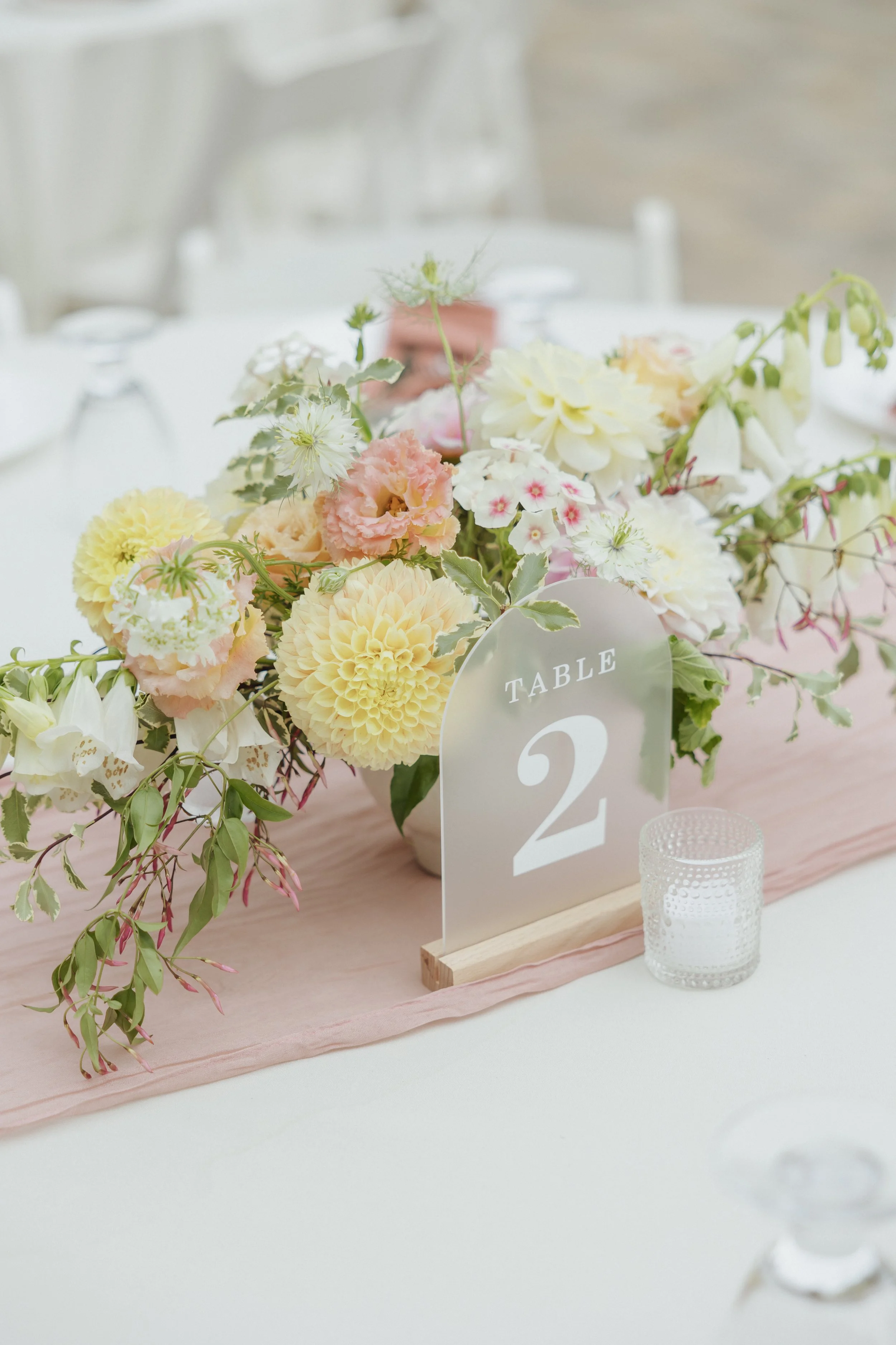 A floral centerpiece with pink, white, and yellow flowers on a table, with a table number card labeled '2' and a small textured glass candle holder.