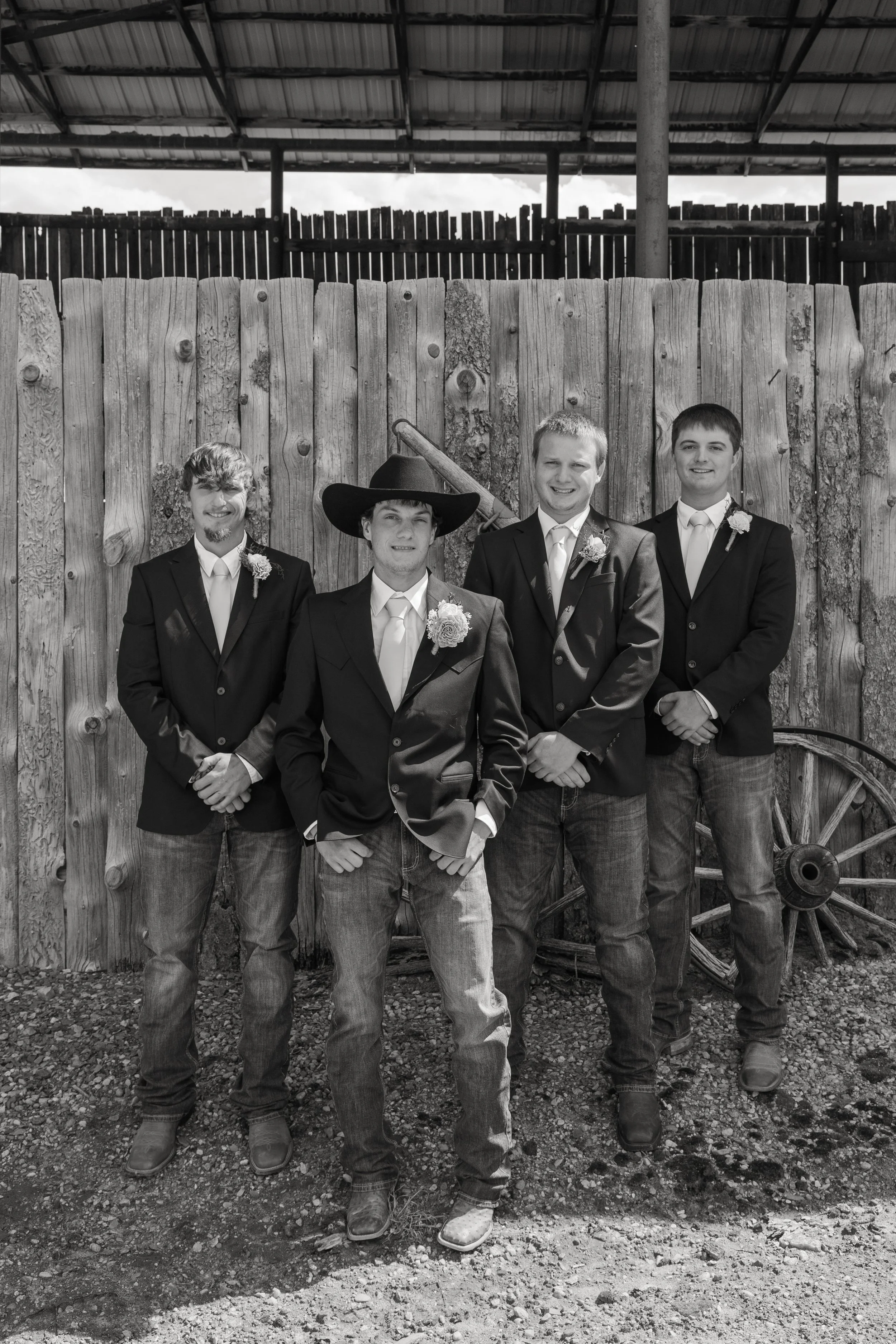 Four men standing together outdoors in front of a wooden fence and rustic wagon wheel, dressed in suits and smiling for a photo, with a barn or shed roof above and cloudy sky in the background.
