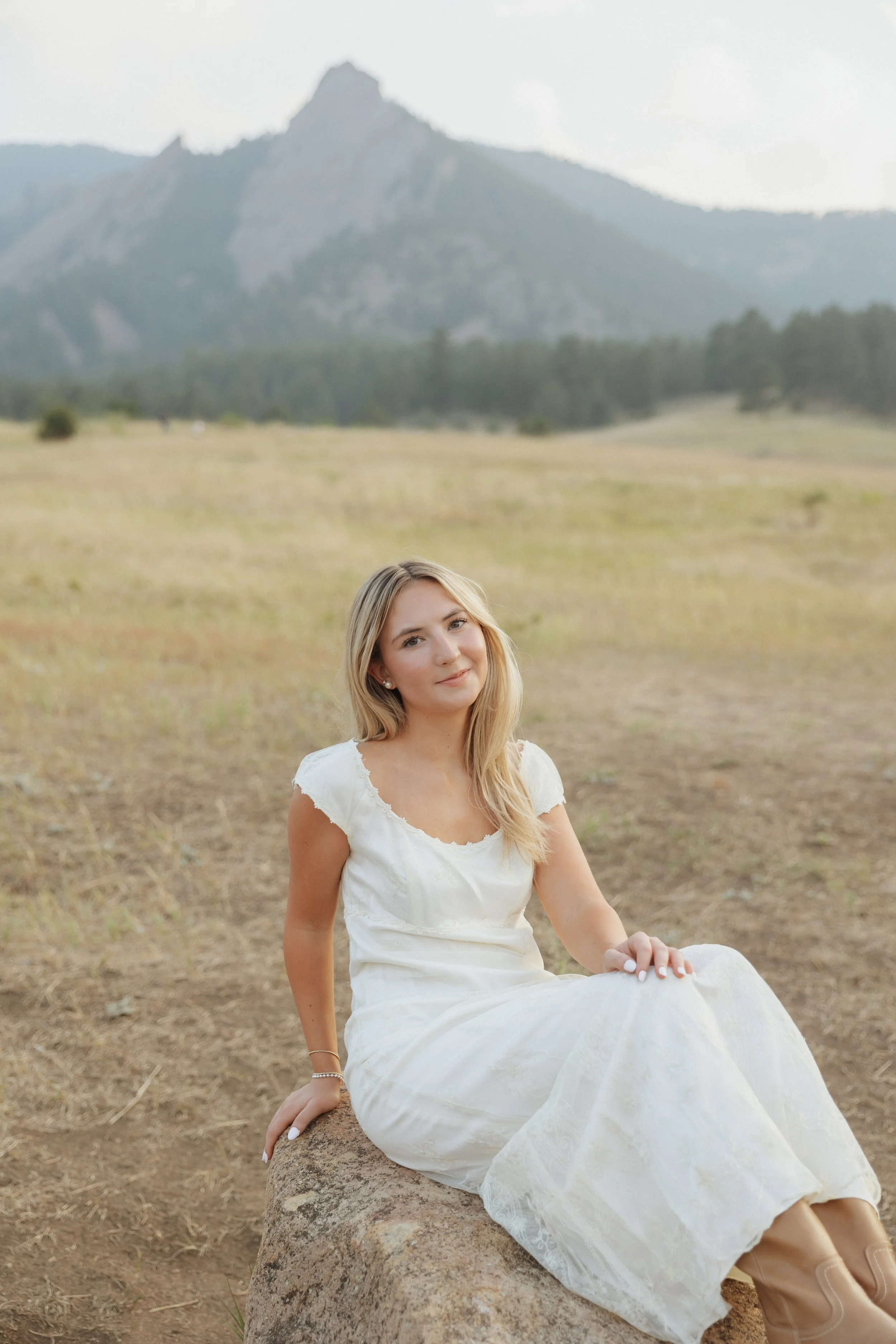 A young woman with blonde hair sitting on a large rock in an open field with mountains in the background, wearing a white dress and looking at the camera.