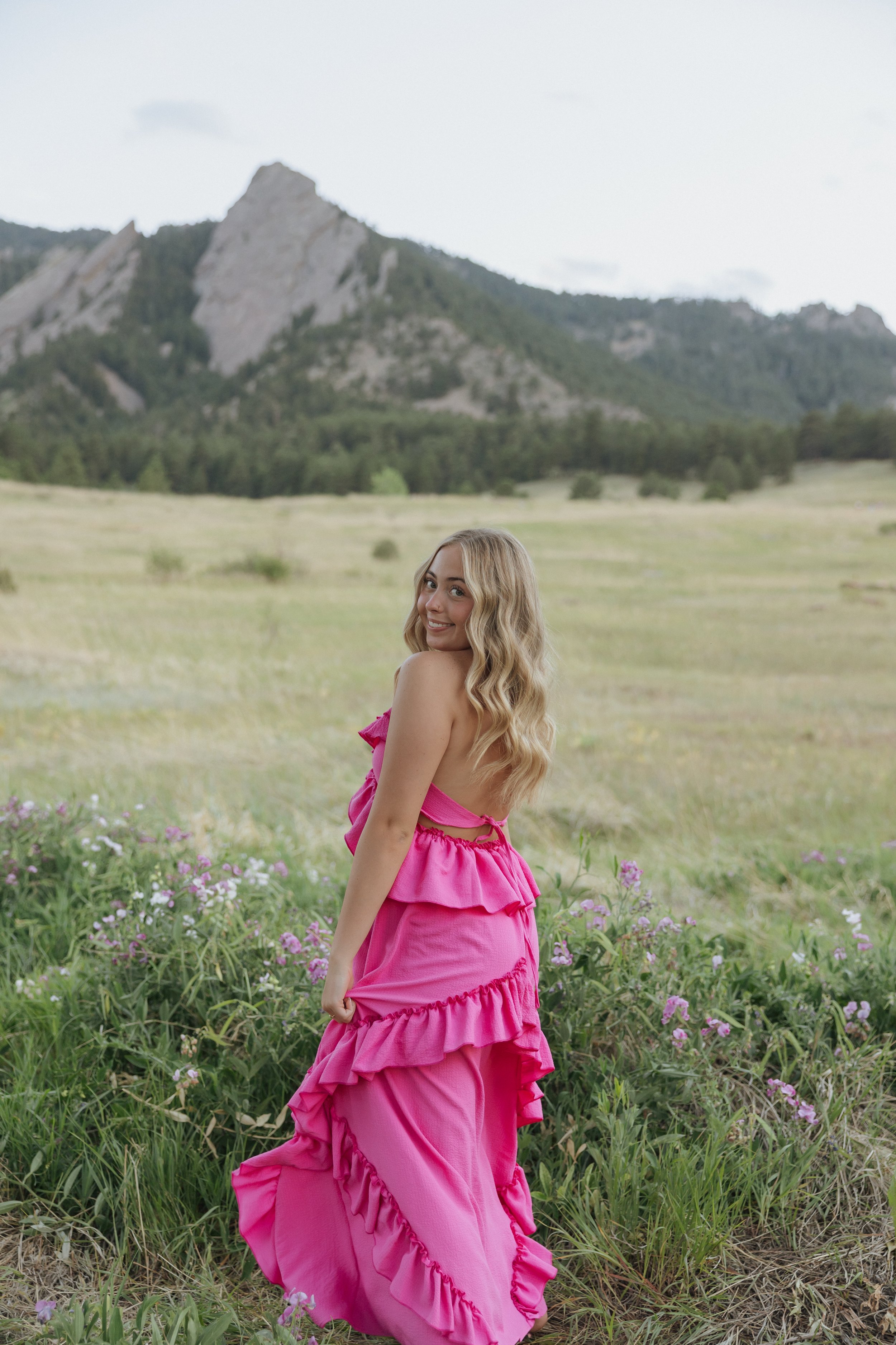 A woman in a bright pink dress standing in a field of green grass and pink flowers, with mountains in the background under a cloudy sky.